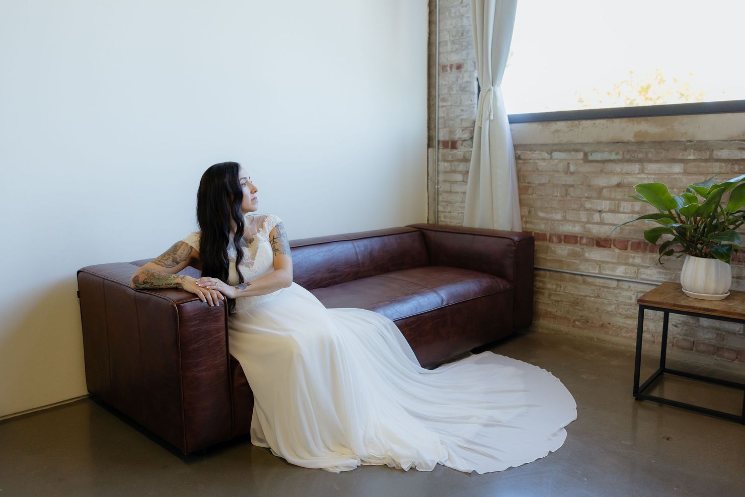 Bride in white wedding gown sitting on brown leather sofa near brick wall and window with curtains.