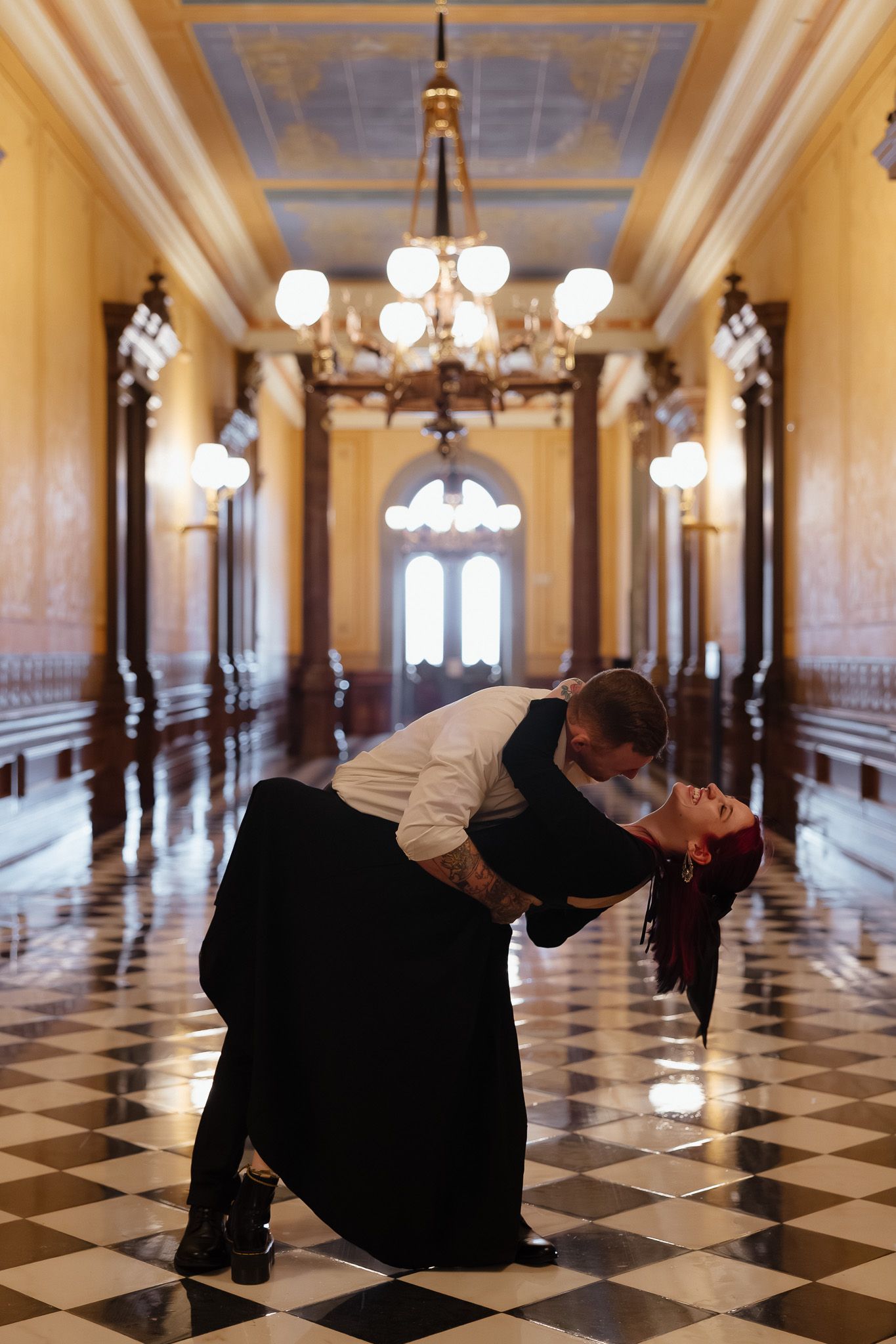 Couple sharing a romantic dip kiss in an elegant grand hallway with chandeliers and marble floors.