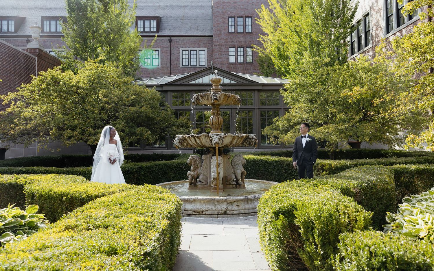 Bride and groom stand apart near an ornate stone fountain in a lush garden courtyard of a brick building.