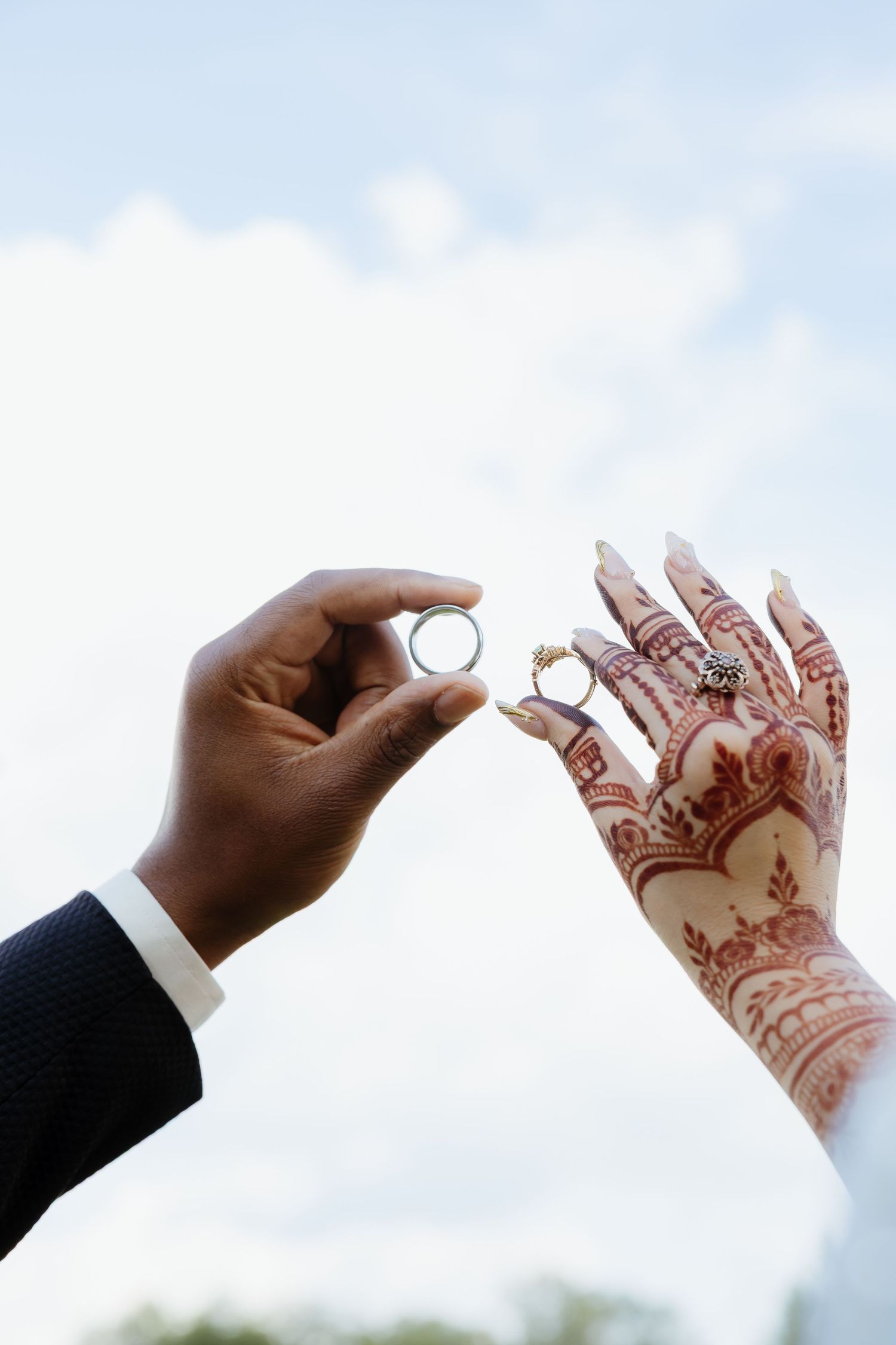 Groom holding wedding ring near bride's henna-adorned hand with rings against a cloudy sky backdrop.