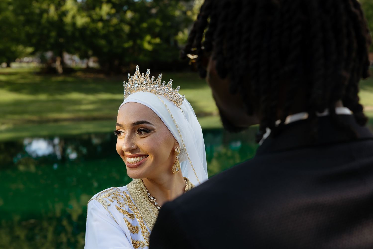 Smiling bride in white hijab and gold crown stands outdoors near pond with groom in black suit.