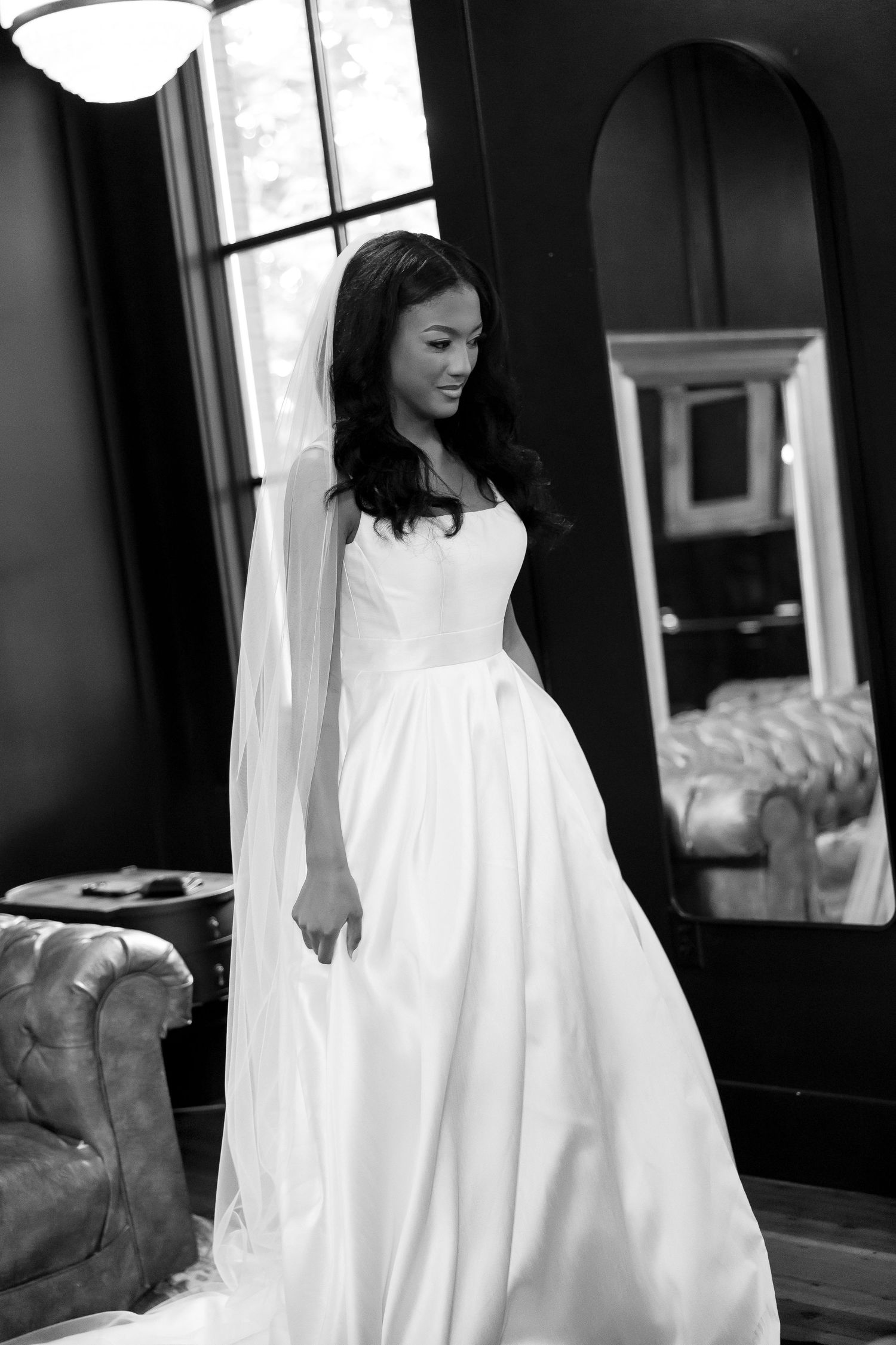 Black and white photo of a bride in a flowing white gown and veil, standing elegantly in a sophisticated room.