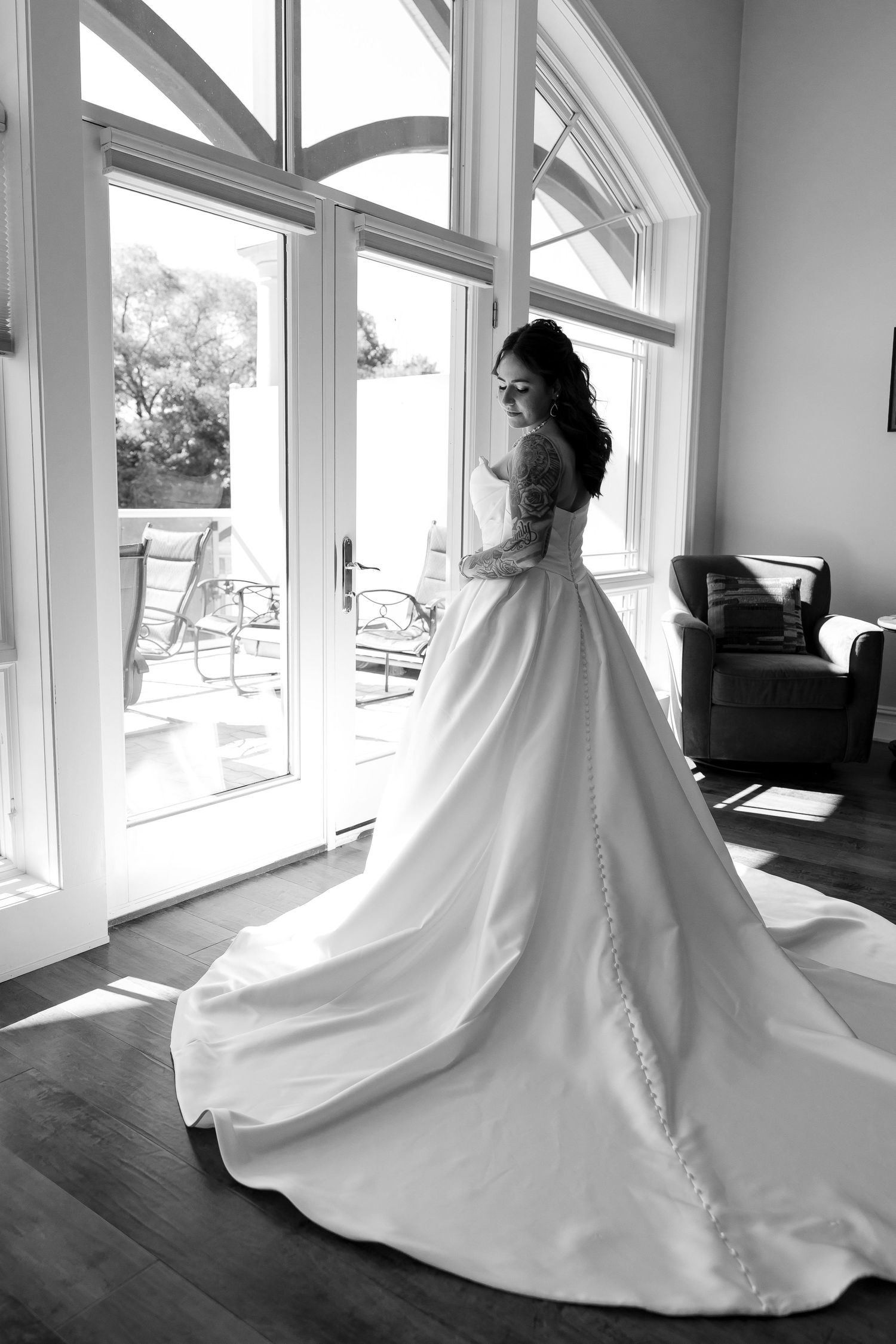 Black and white photo of a bride in a flowing ball gown with a long train standing by large windows.