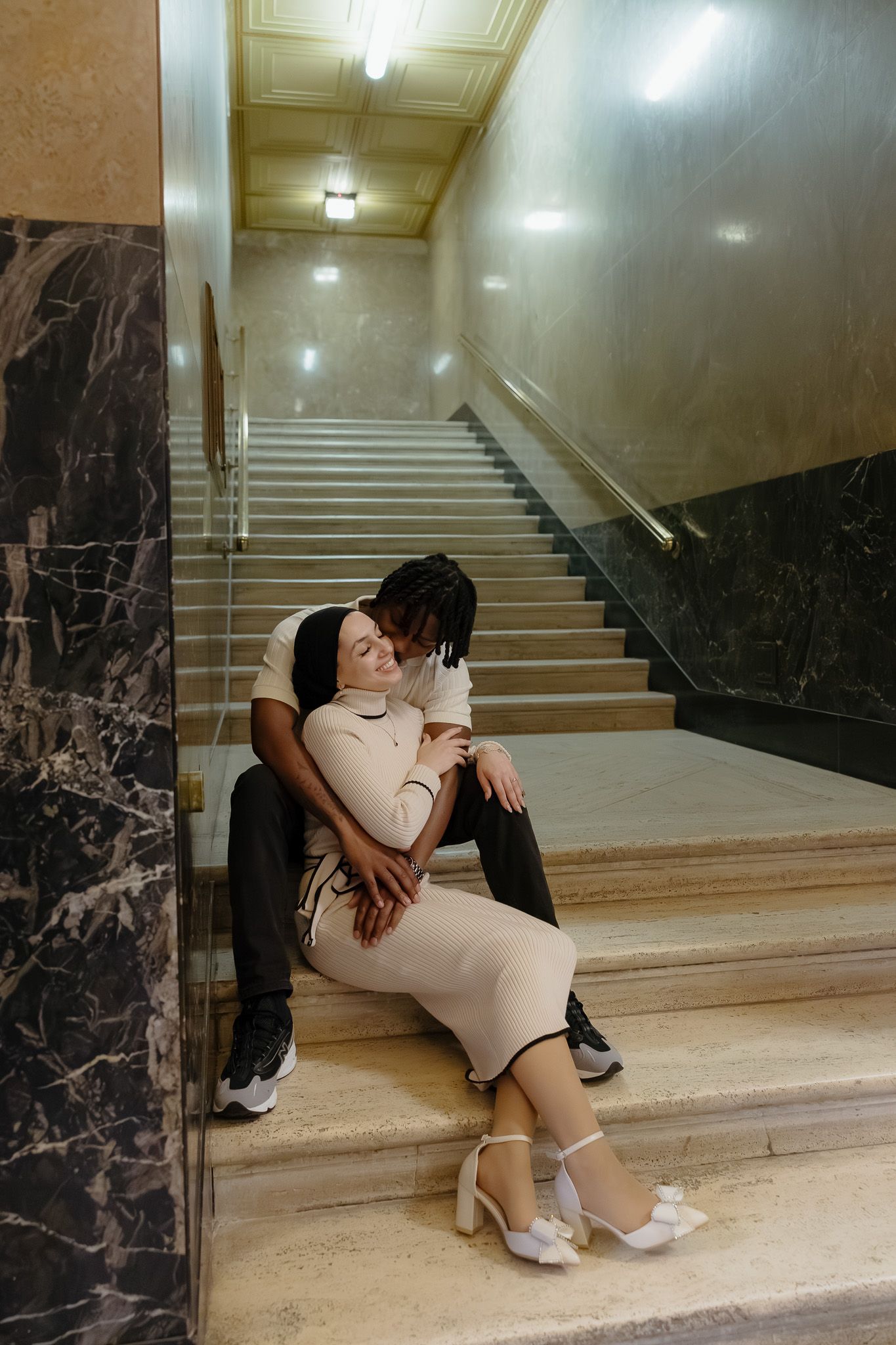 Couple sitting on marble stairs in elegant building, woman in white dress leaning on man in black outfit.