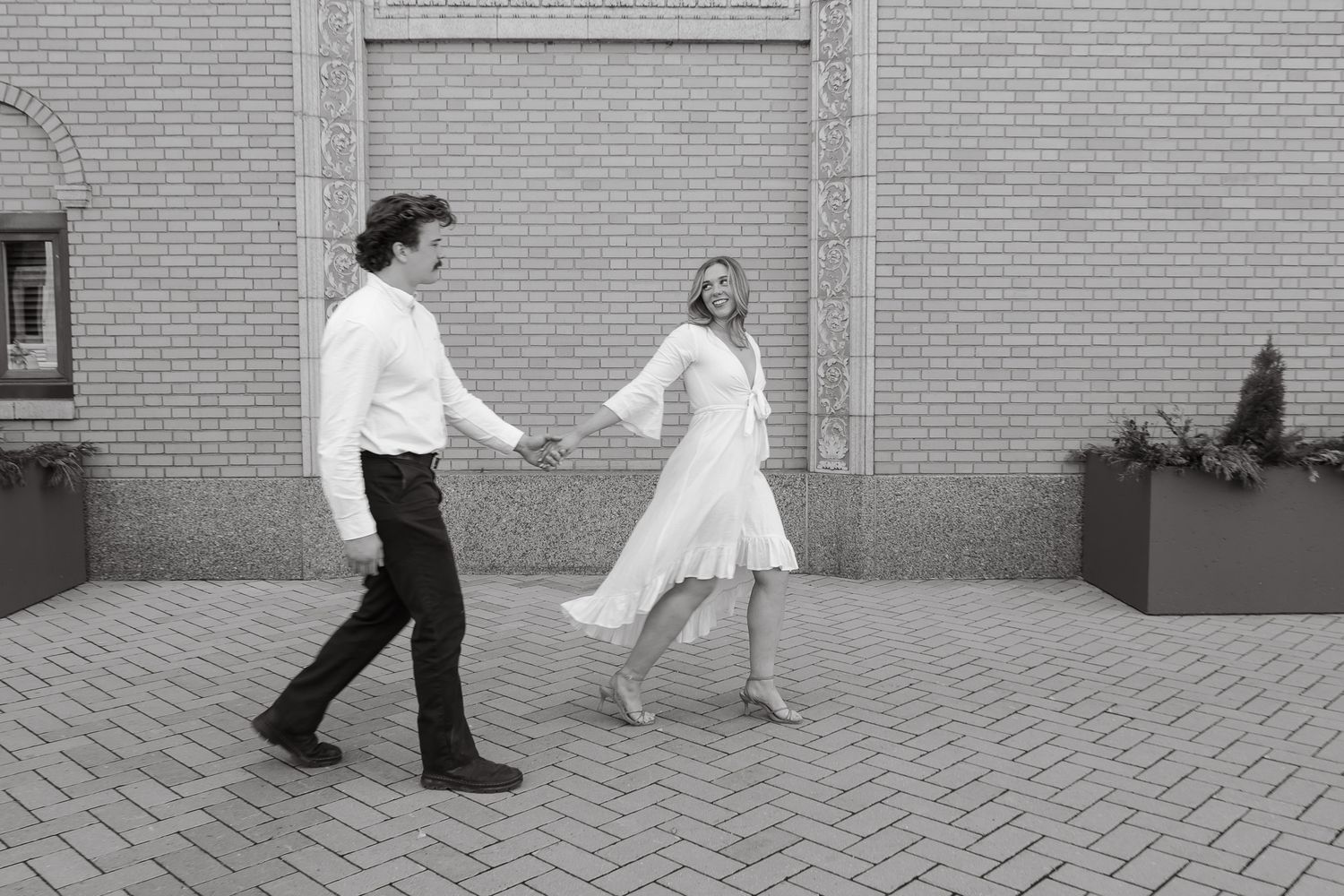 Black and white photo of a couple holding hands and dancing outdoors on a brick plaza near a building.