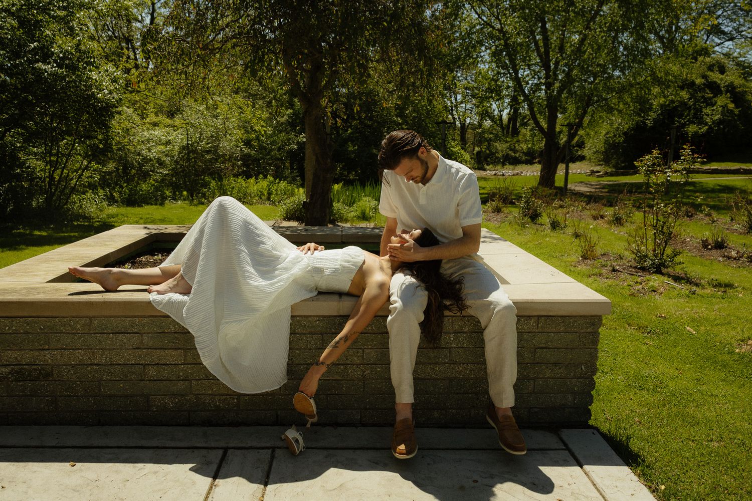 Couple in white outfits sharing a romantic moment on a stone wall in a lush green park on a sunny day.