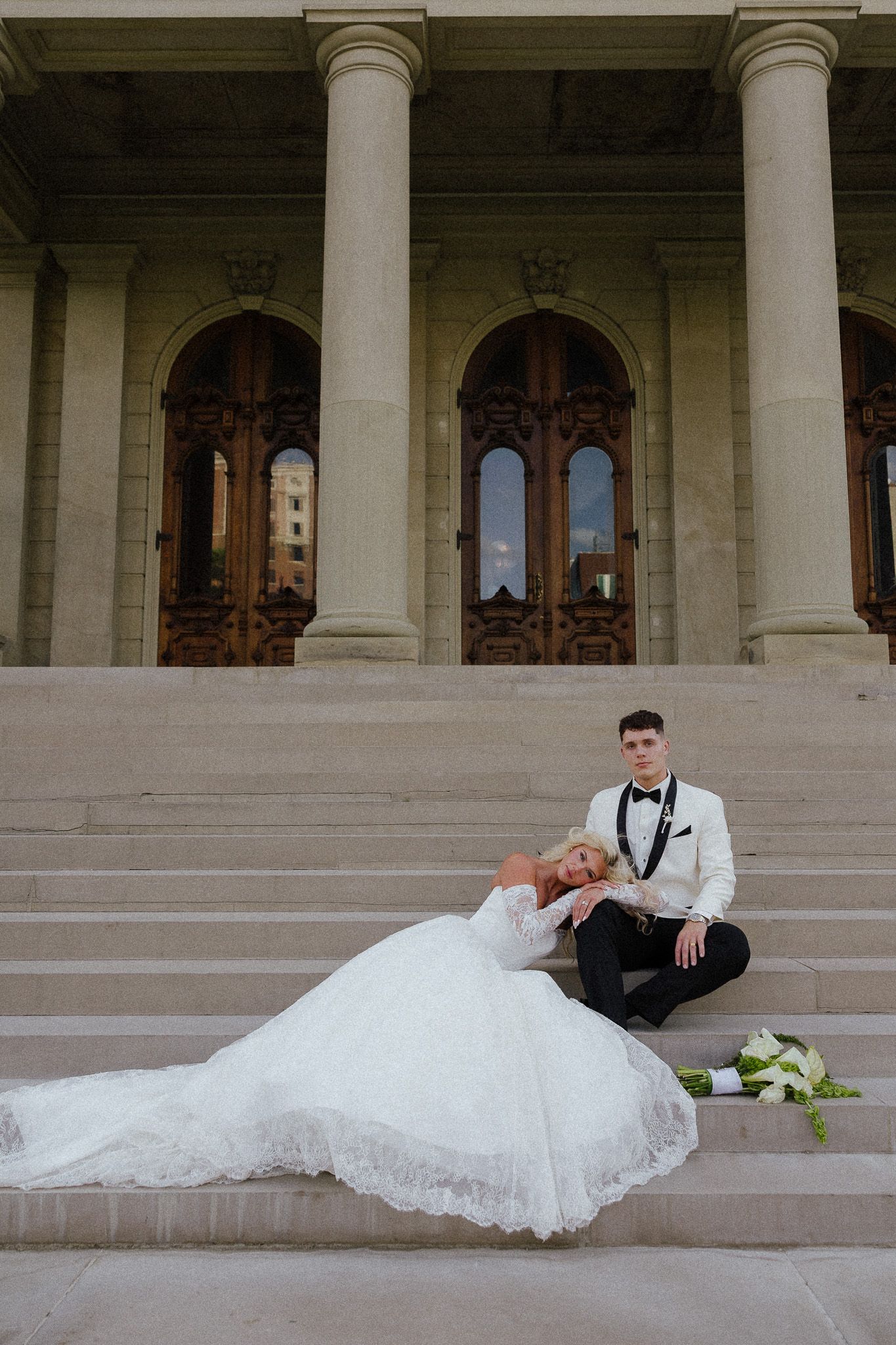 Bride in white lace gown reclines on stone steps beside groom in white tuxedo at elegant columned building.