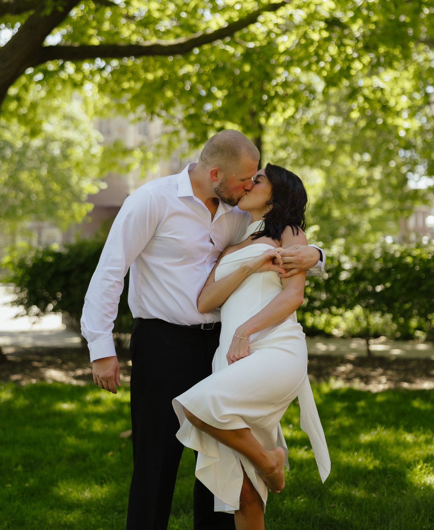A couple shares a romantic kiss outdoors, the woman in a white dress and man in white shirt, surrounded by lush green trees.