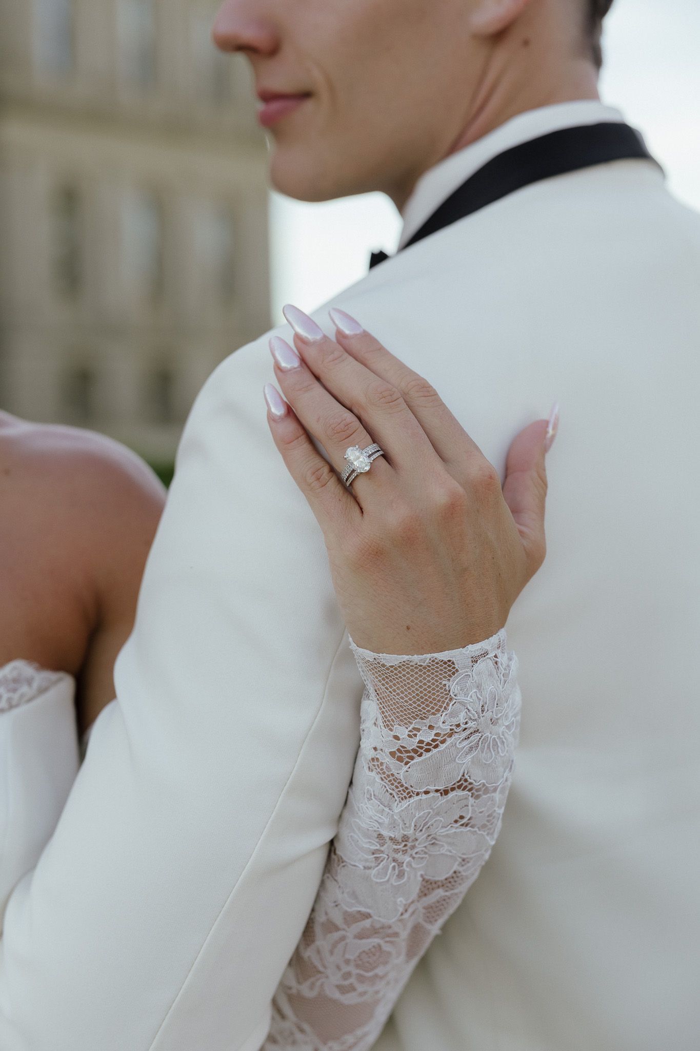 Bride in lace wedding dress showing diamond engagement ring on groom's white tuxedo jacket.