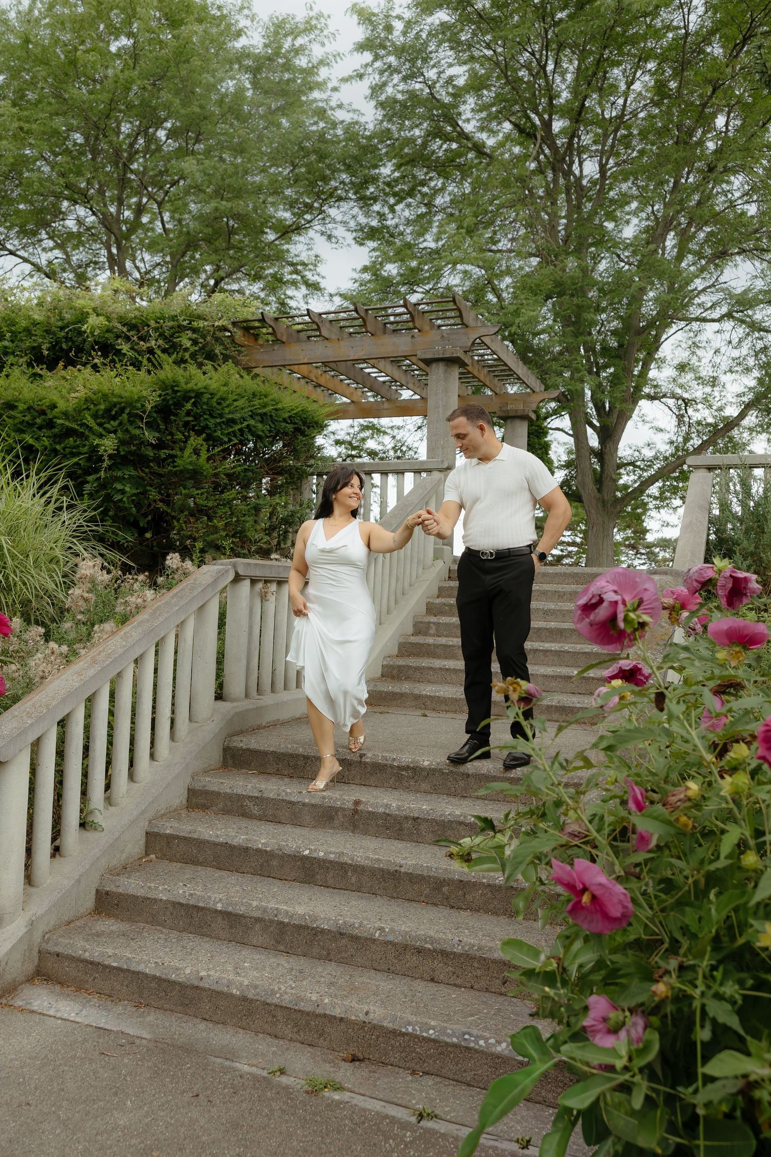 Couple holding hands walking down stone steps in a garden with pink roses and green trees.