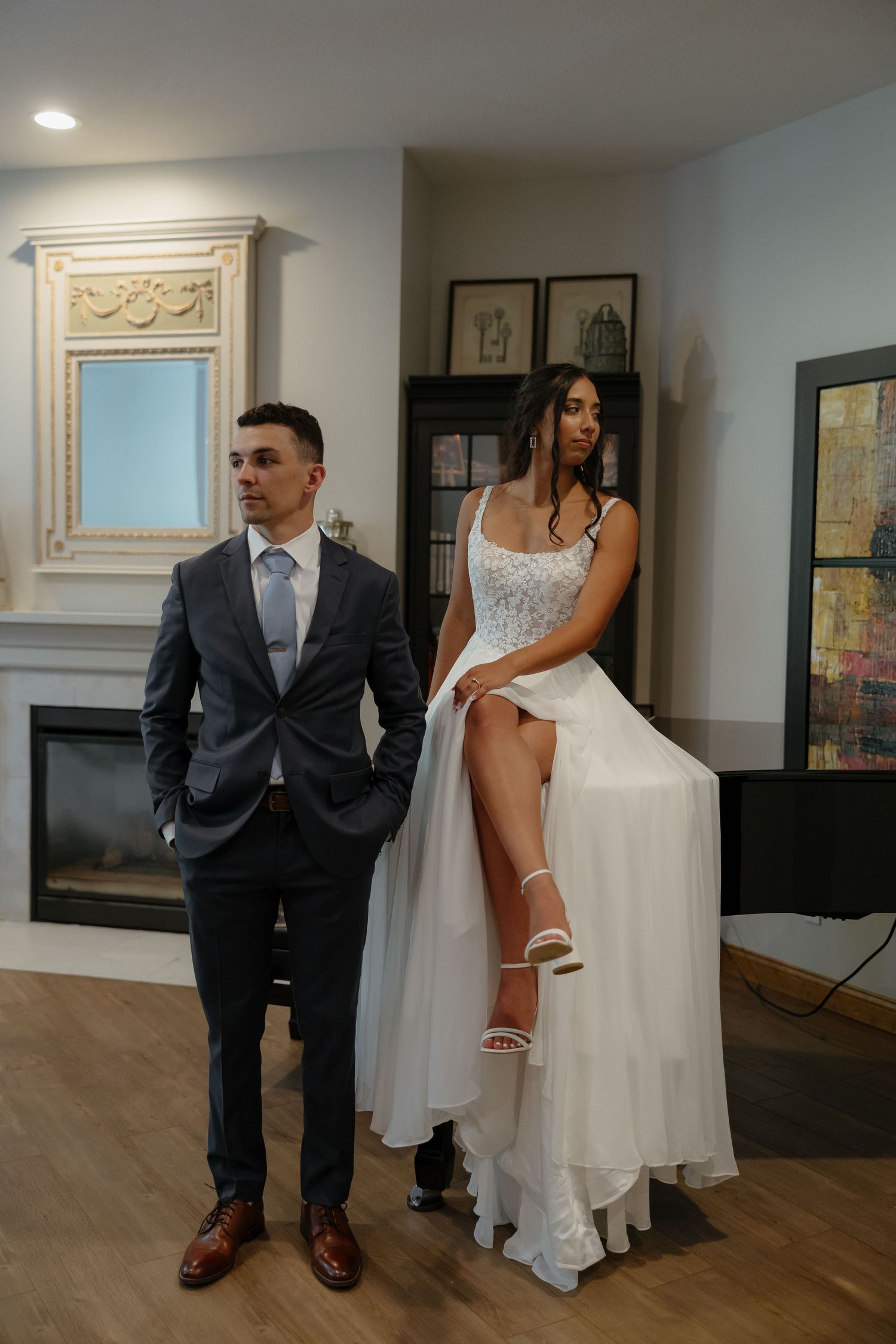 Bride in white lace gown with slit and groom in dark suit pose together indoors near a fireplace.