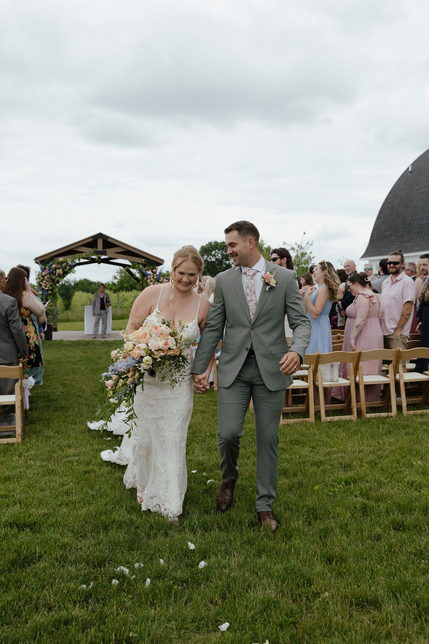 Bride and groom walk down outdoor aisle after wedding ceremony at rustic barn venue on cloudy day.