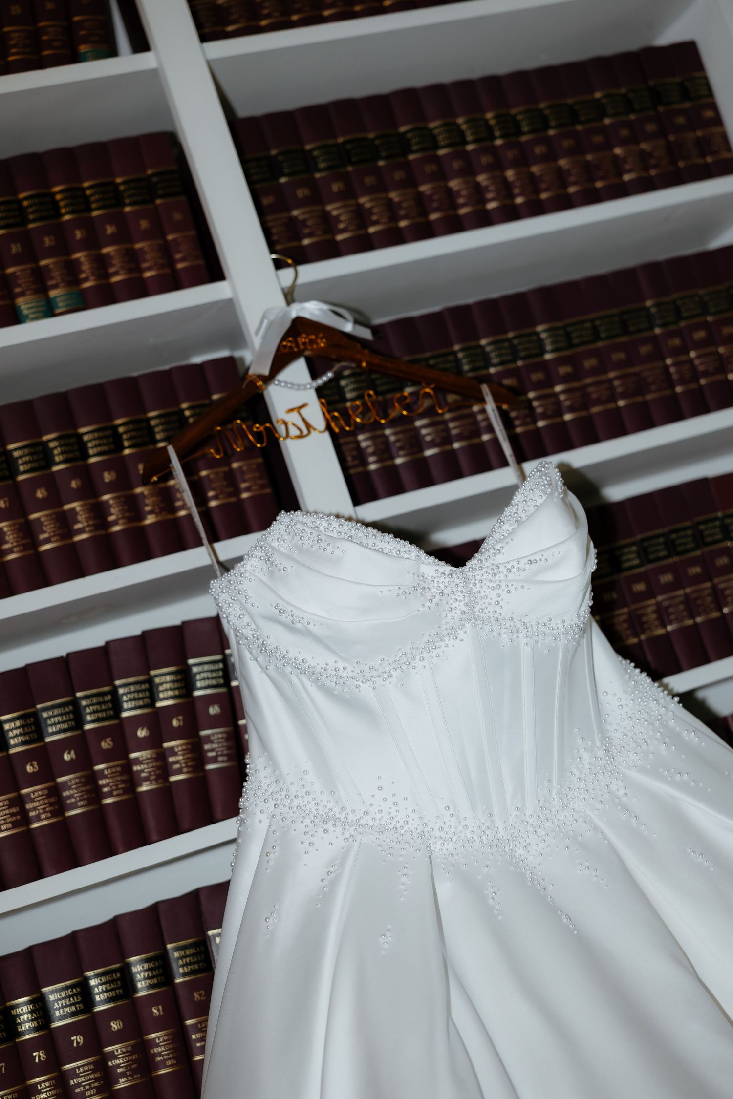 White beaded strapless wedding dress hanging on a personalized wooden hanger in front of a bookshelf filled with books.