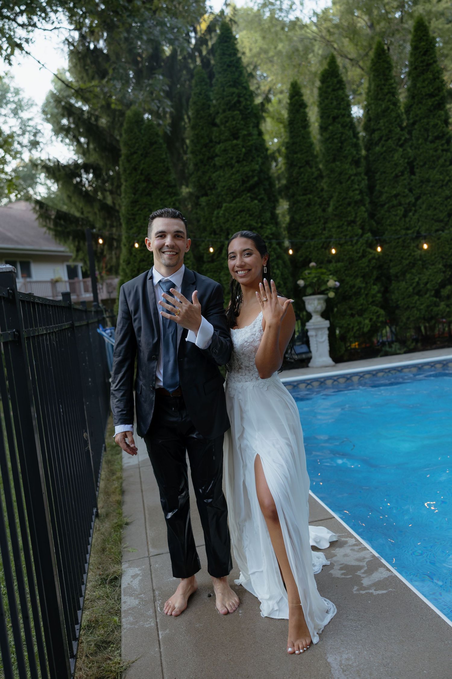 Newlyweds in wedding attire show off rings poolside, groom in suit and bride in white gown with slit.