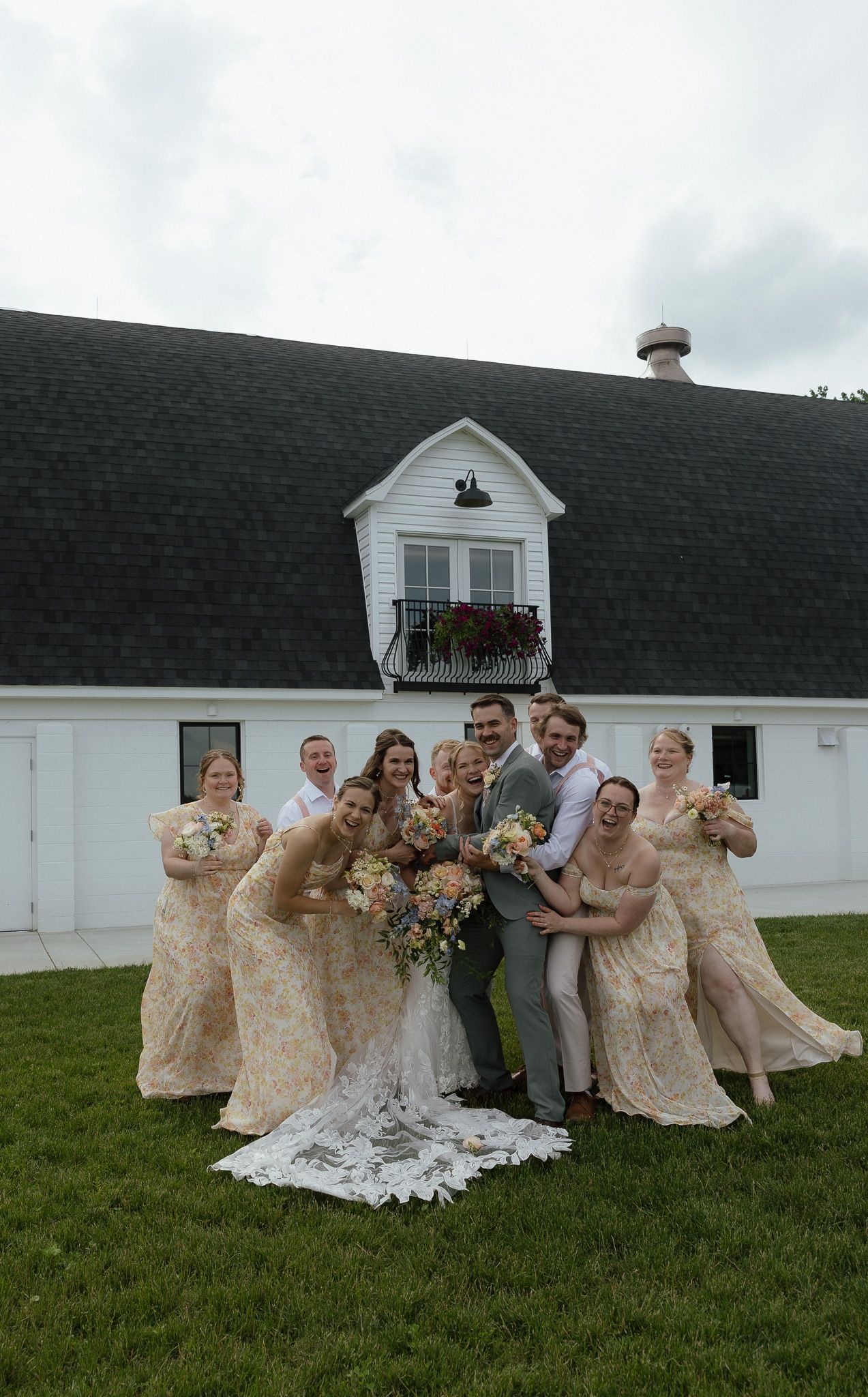 Wedding party poses joyfully outside white barn venue with bridesmaids in floral dresses.