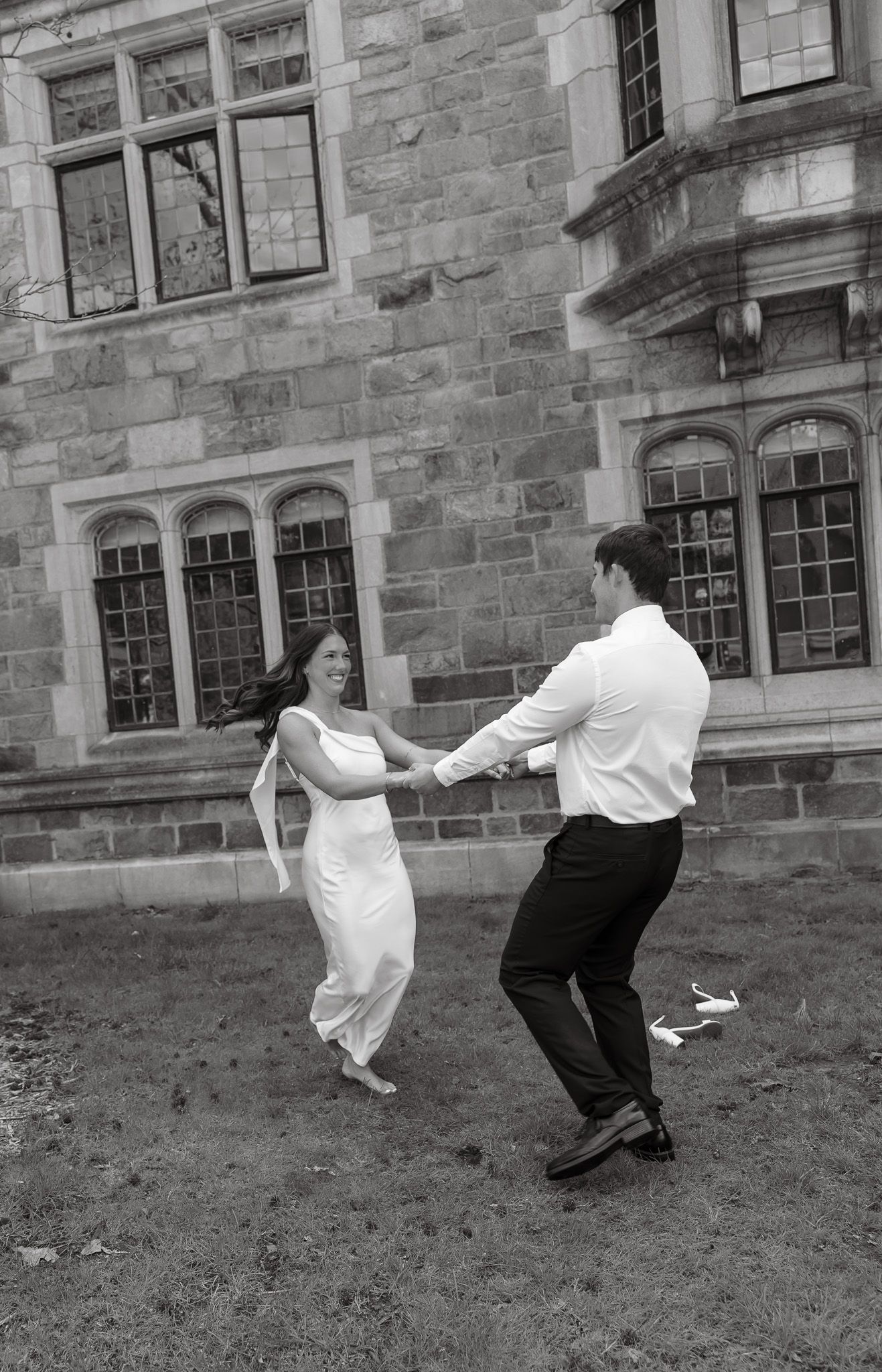Black and white photo of a couple dancing joyfully outside a stone building, bride in white dress, groom in formal attire.