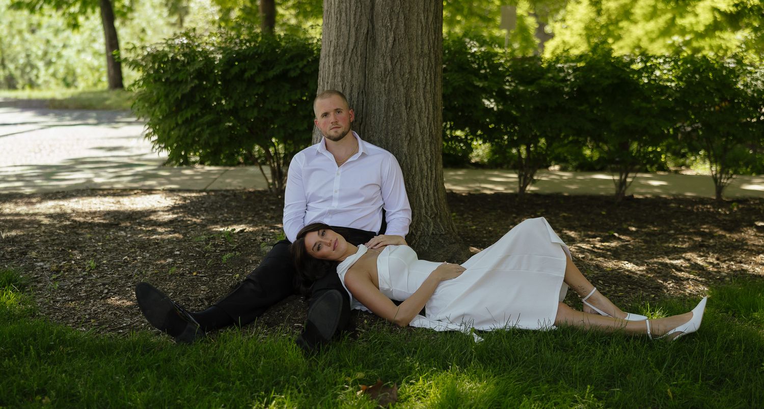 Couple resting against a tree trunk on green grass in a park, woman lying across mans lap.