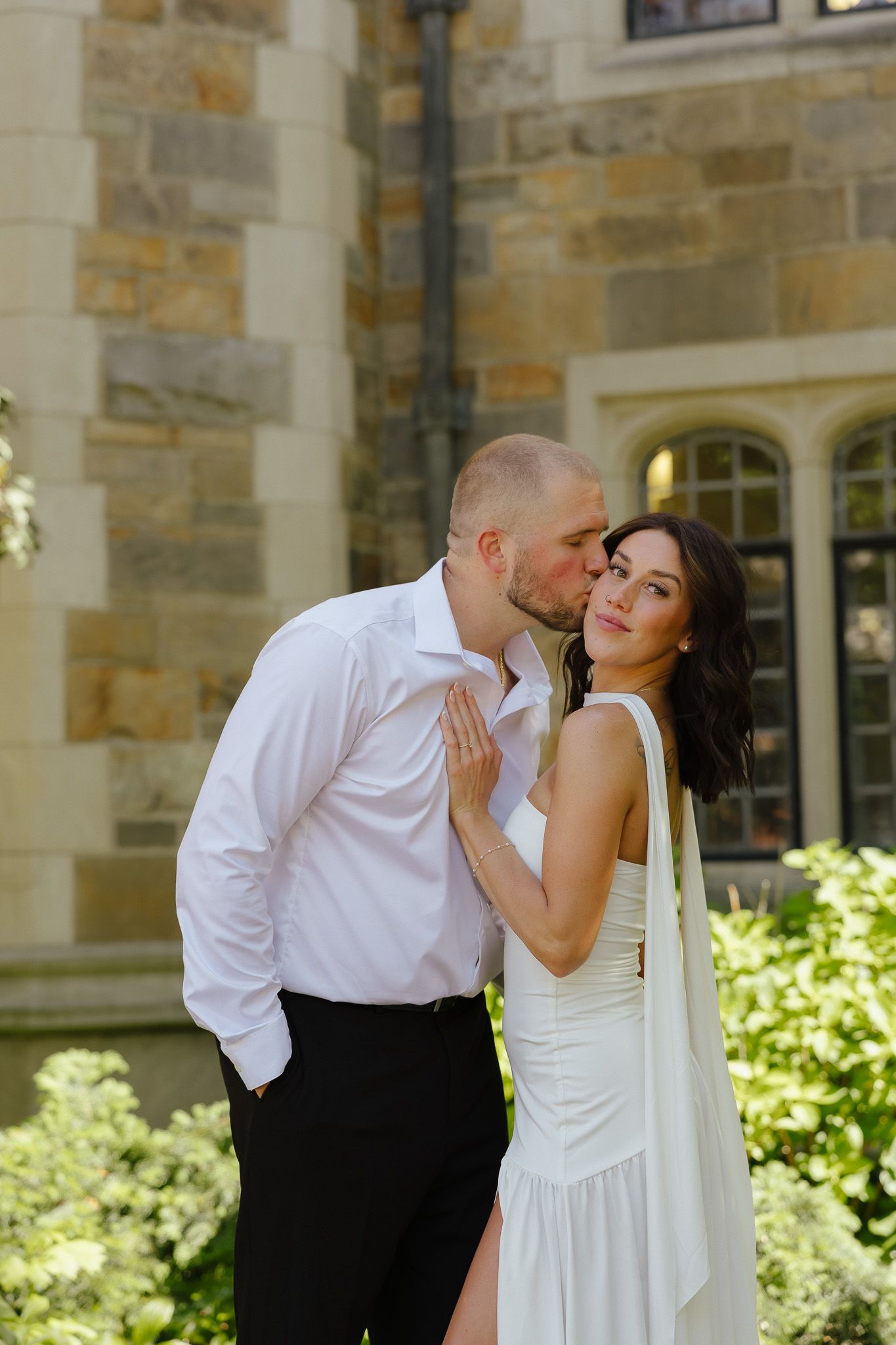 A groom in a white shirt kisses his bride on the cheek as she smiles, posing before a stone building backdrop.