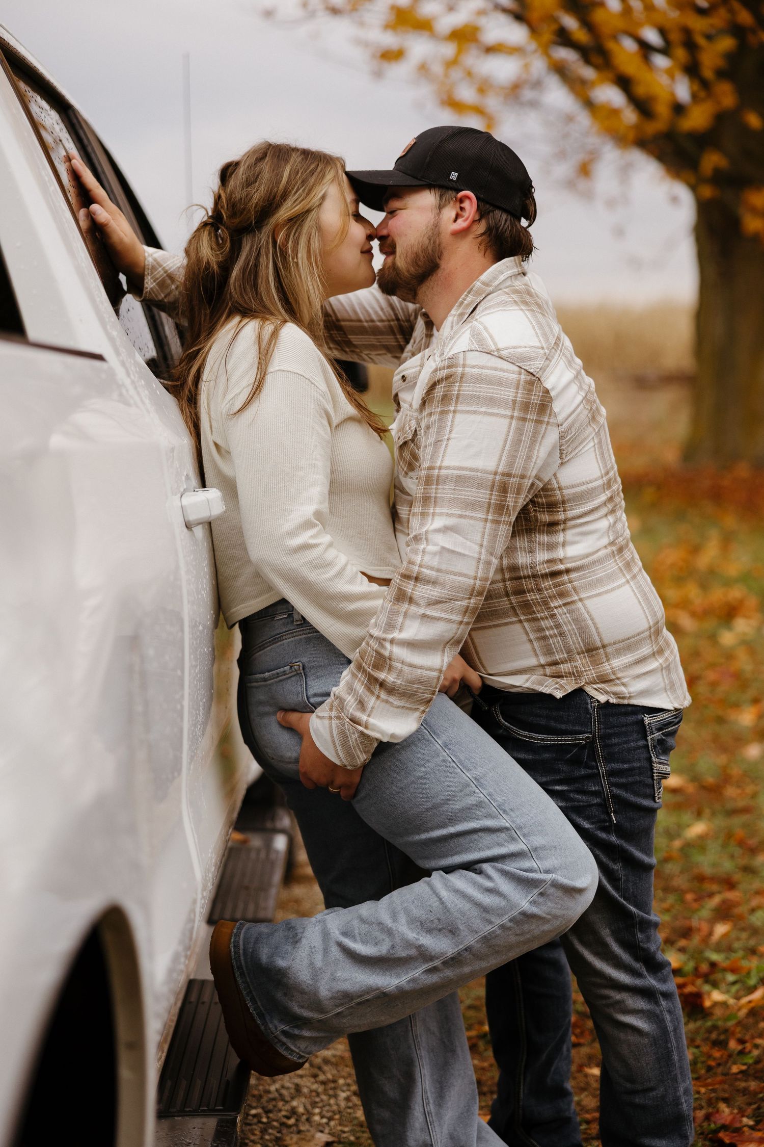A playful engagement photo against a white vehicle with autumn foliage in the background.