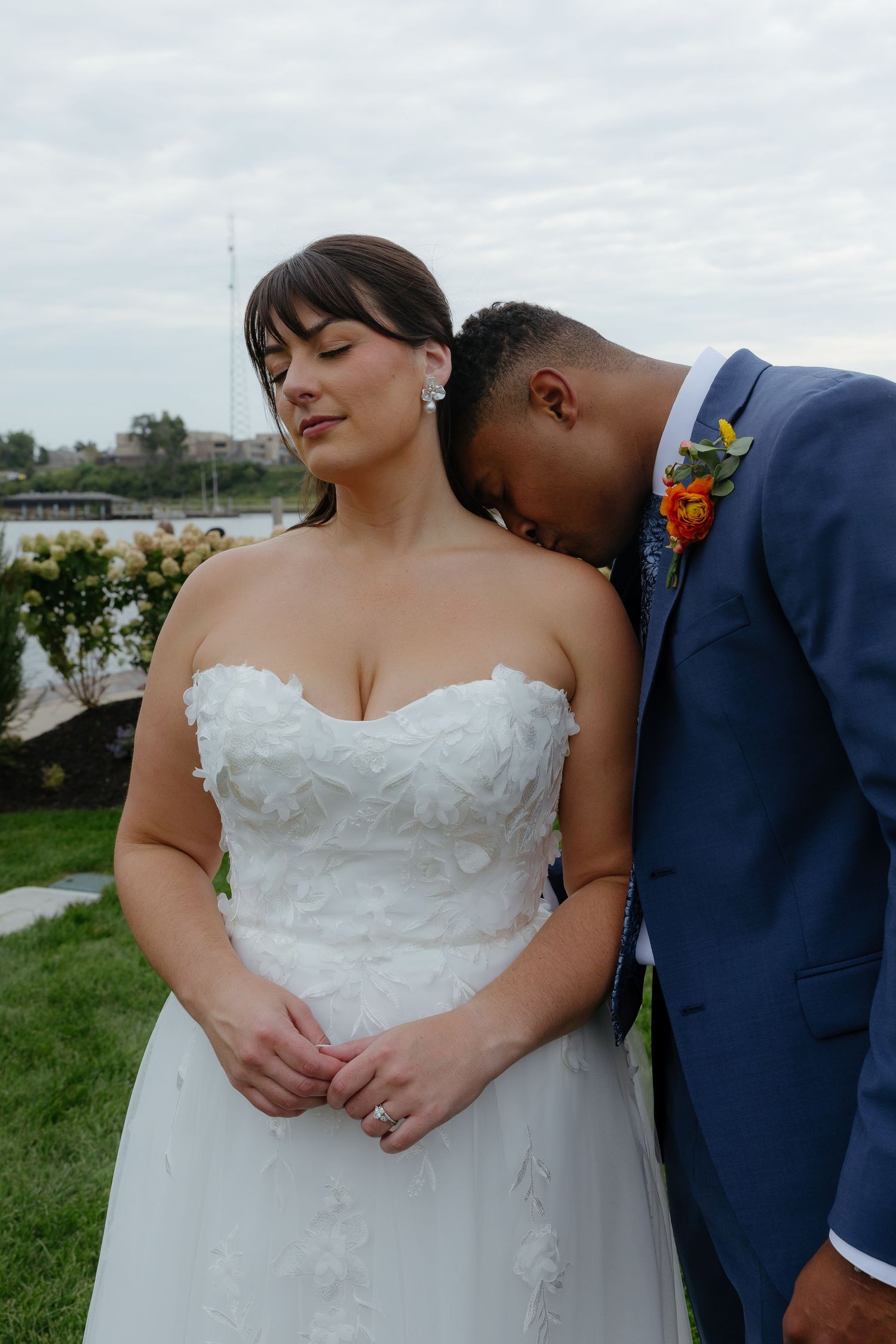 Bride in strapless white gown and groom in blue suit share tender moment outdoors near waterfront.