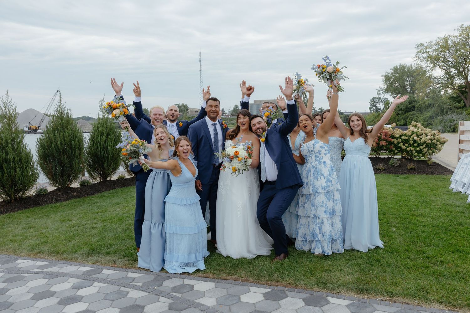 Wedding party in blue dresses and suits celebrating outdoors, raising hands joyfully with bride and groom at center.