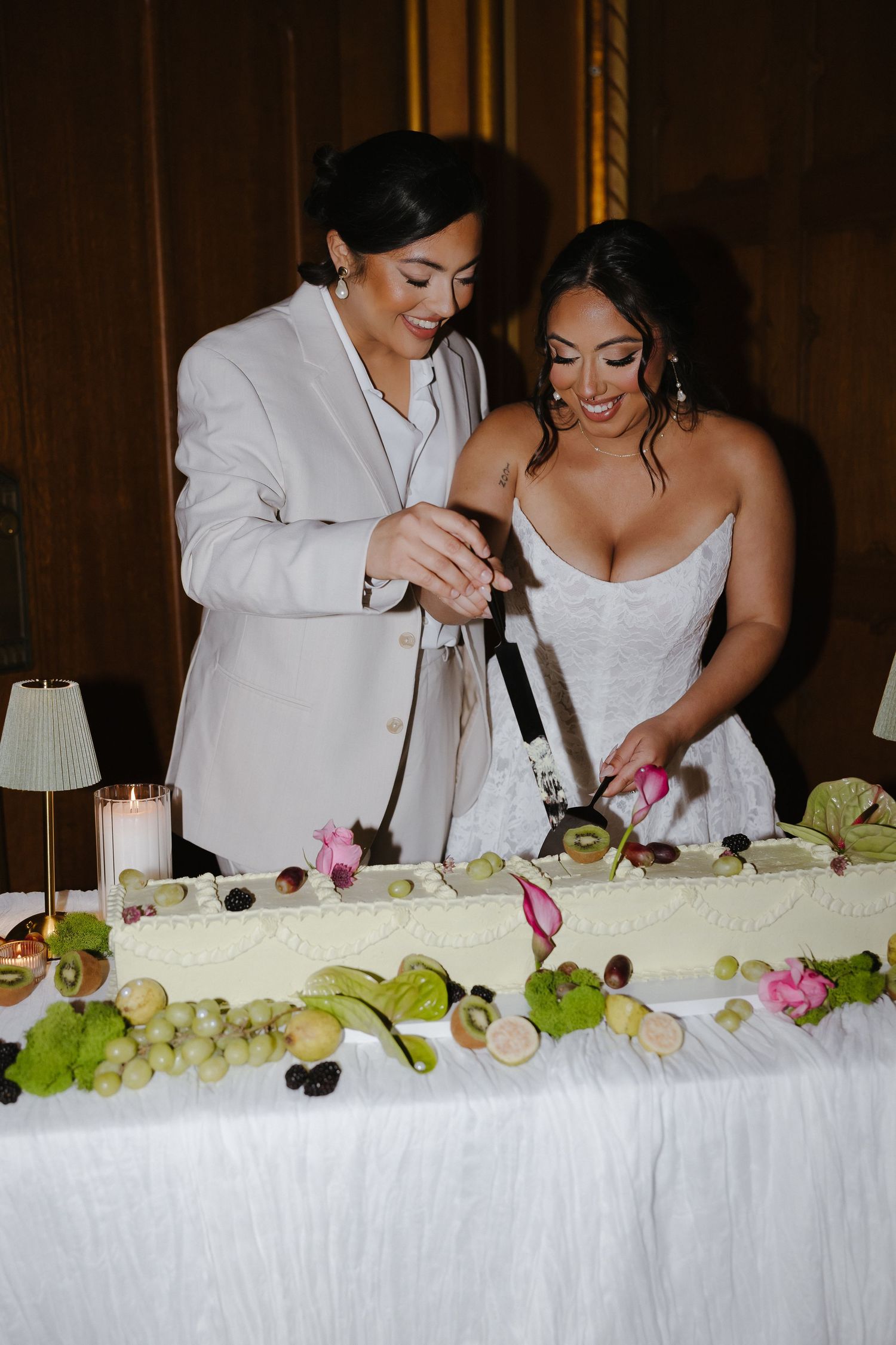 Two brides cutting a elegant white wedding cake decorated with fresh fruit including grapes and kiwi at their reception.
