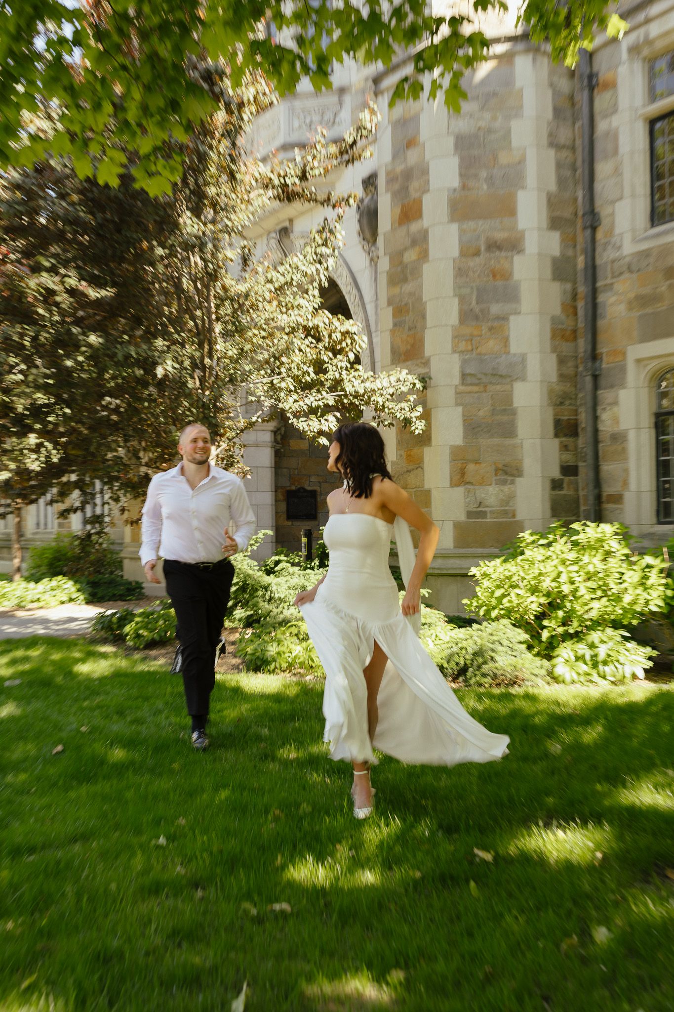 A bride in a white gown and groom in a white shirt run joyfully across green grass near a stone building.