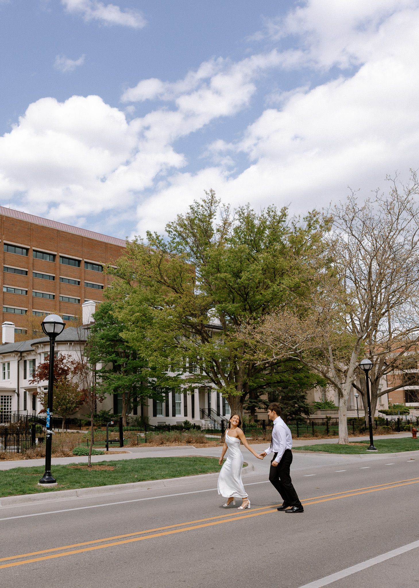 Bride and groom cross a sunny street holding hands, with historic buildings and trees in the background.