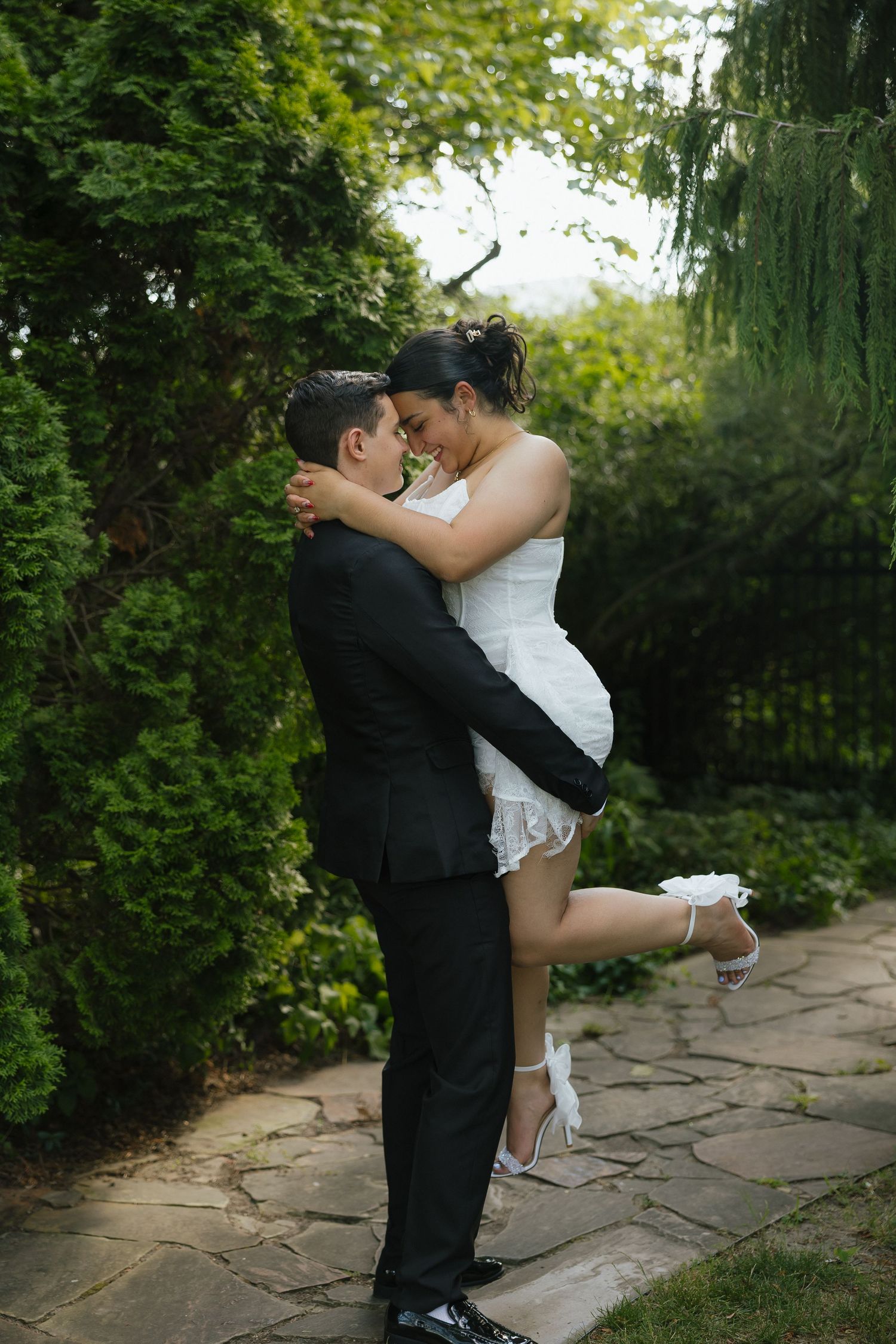 Groom in black suit lifting bride in white dress, sharing romantic moment in lush garden on wedding day.