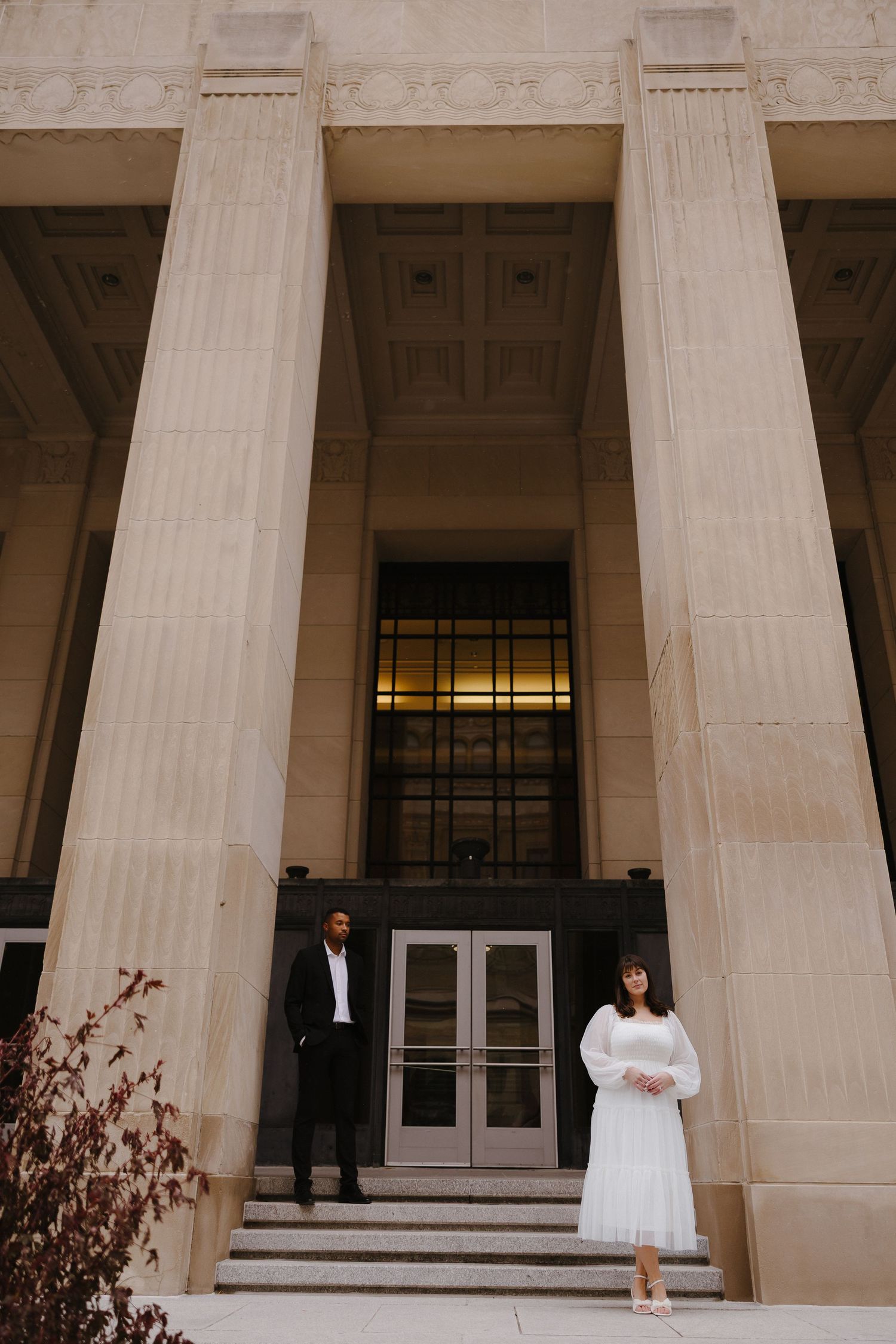 A couple poses dramatically between classical stone columns of a grand building.