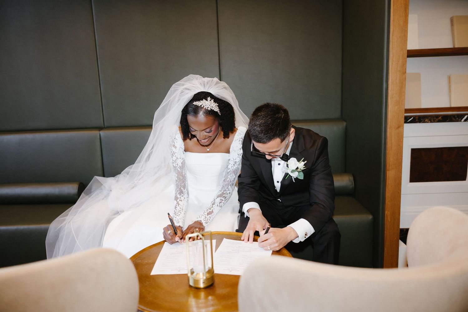 Bride and groom signing wedding documents at a table, dressed in white gown and black tuxedo.