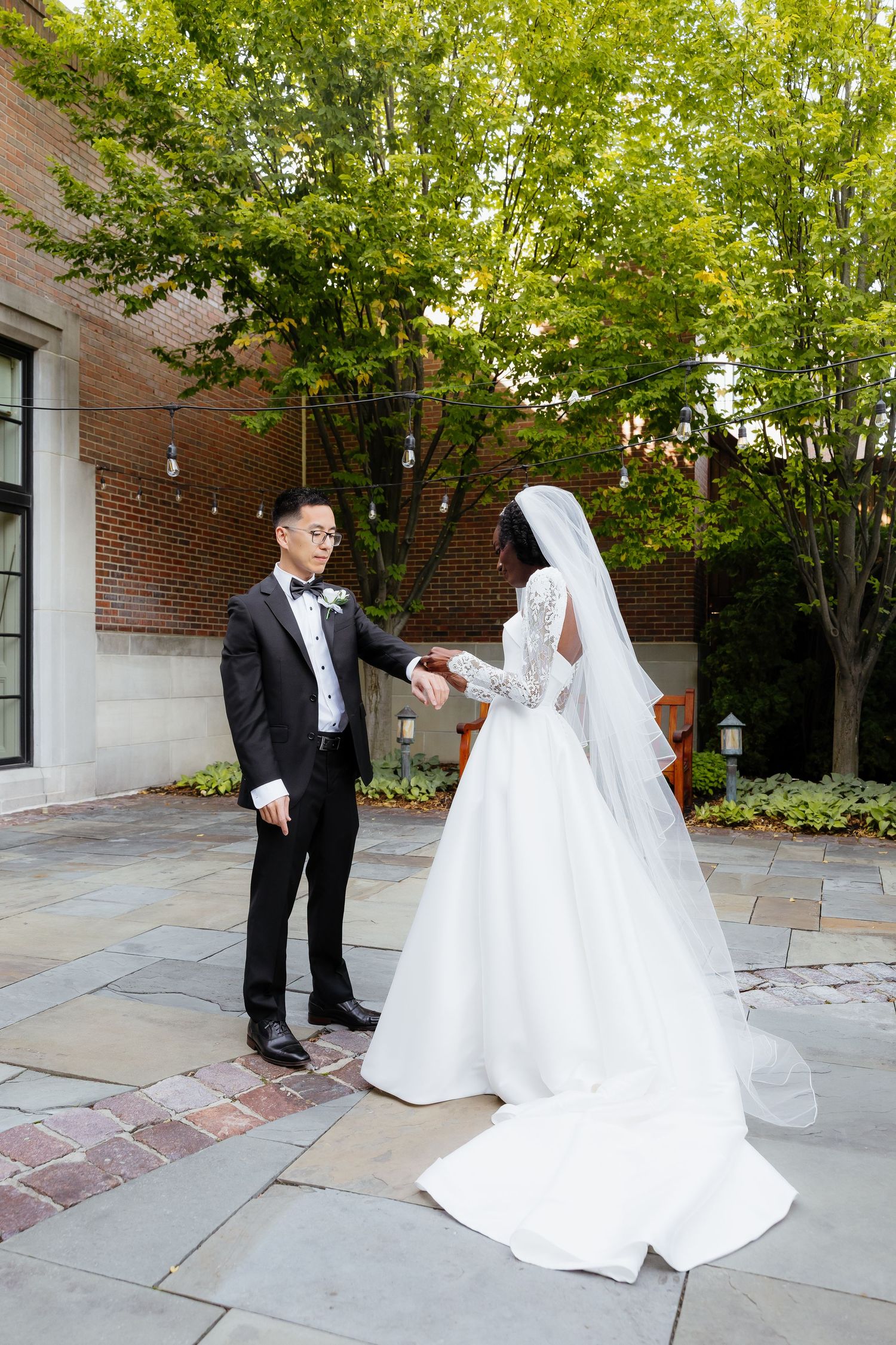 A groom in a black tuxedo holds hands with a bride in a white lace gown and veil in an outdoor courtyard.