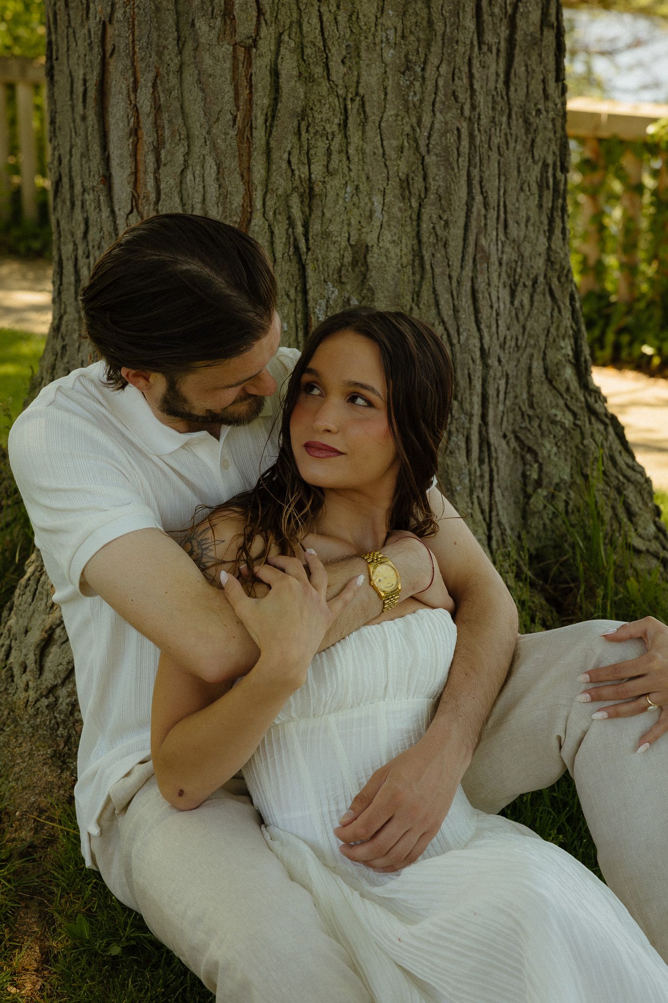 A couple in white outfits sits against a large tree, sharing a tender embrace in a lush green outdoor setting.