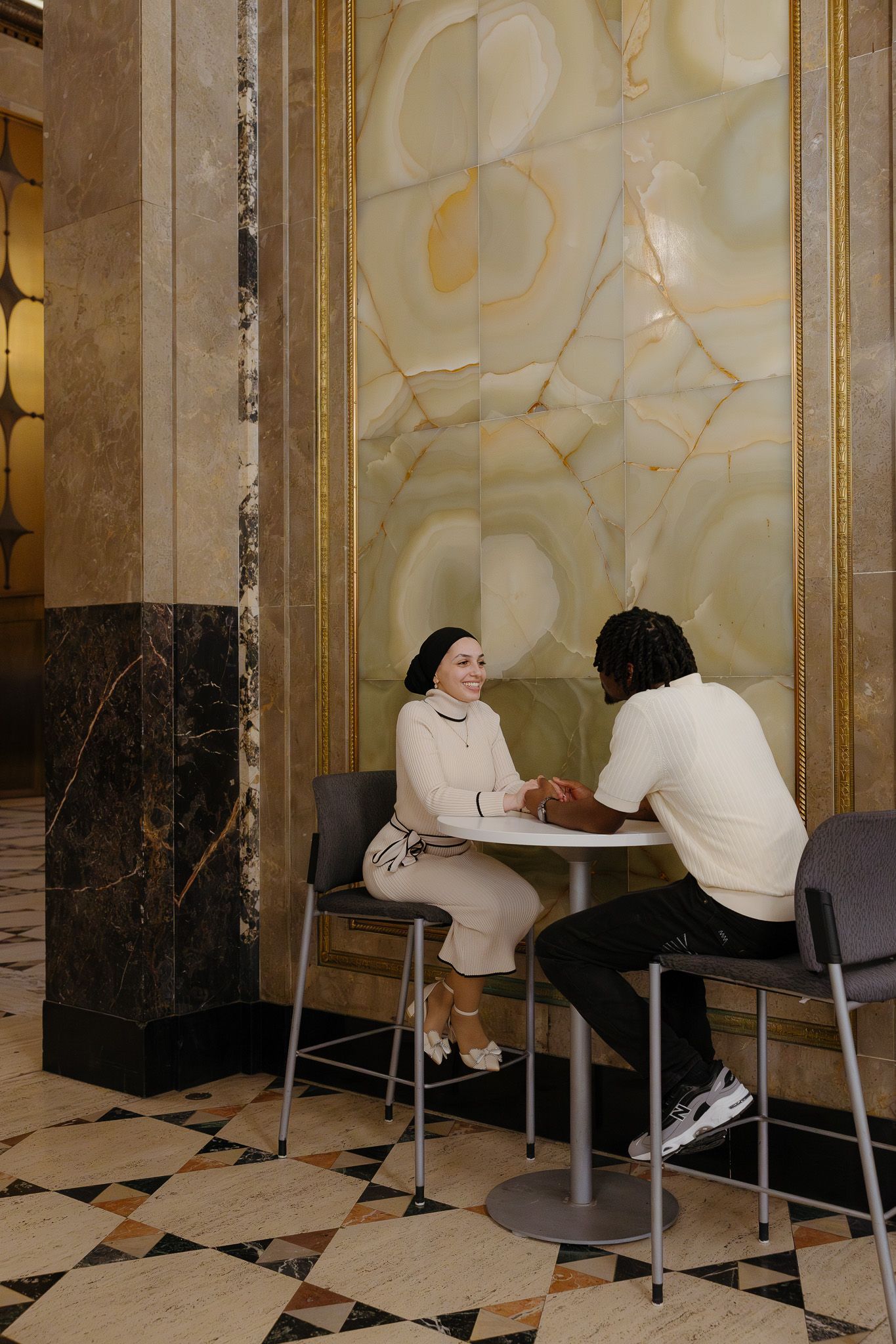 A couple holds hands at a small round table in an elegant marble-walled lobby with decorative tile floors.