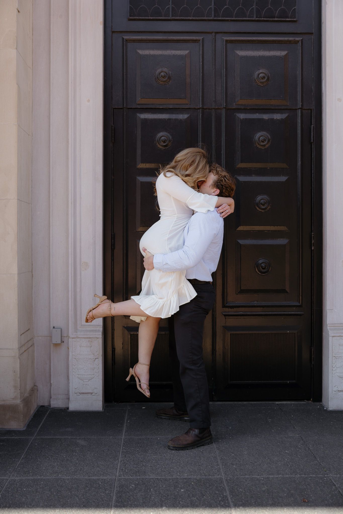 Couple sharing a romantic embrace in front of a grand dark wooden door, man lifting woman in white dress.