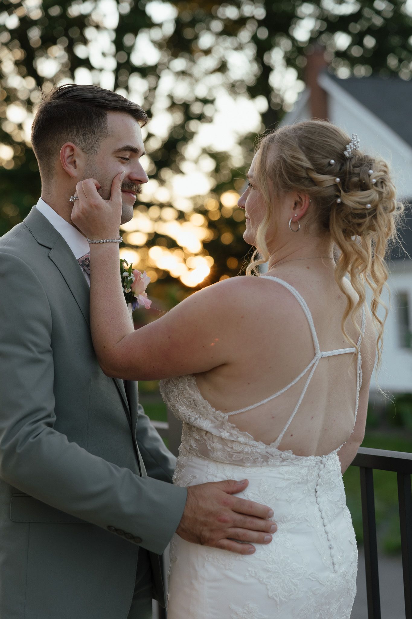 Bride tenderly touches grooms face as they share an intimate moment at sunset, both smiling in wedding attire.
