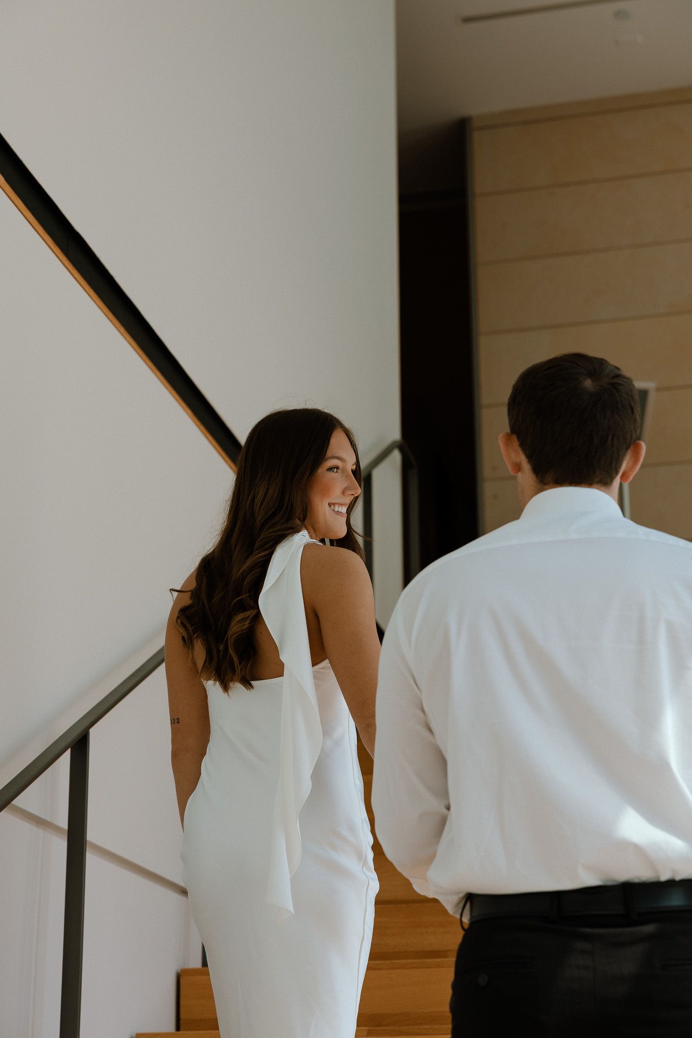 Smiling bride in white backless gown glances over shoulder at groom in white shirt on modern staircase.