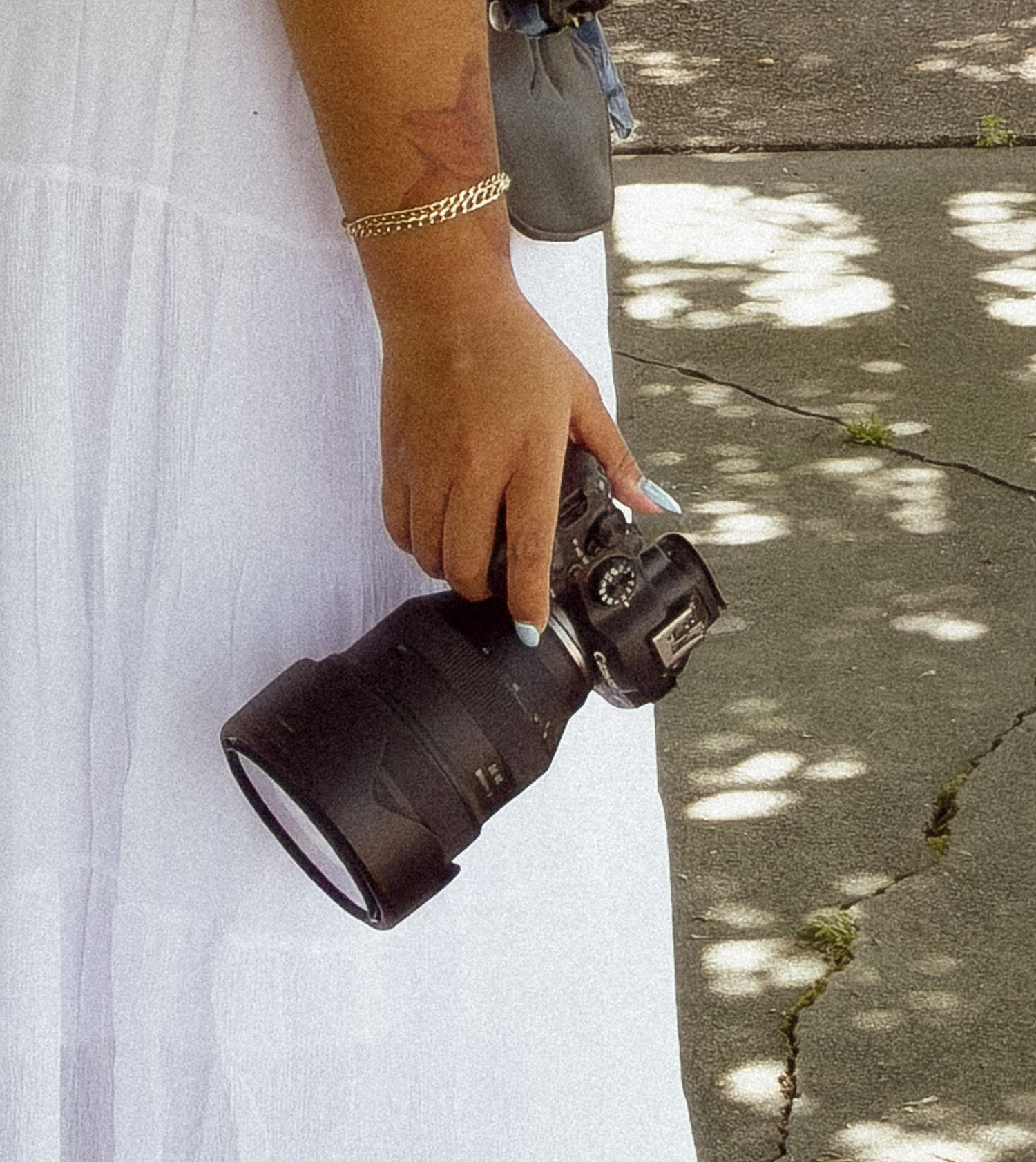 Woman in white dress holding a professional DSLR camera with large lens outdoors, wearing a gold bracelet.