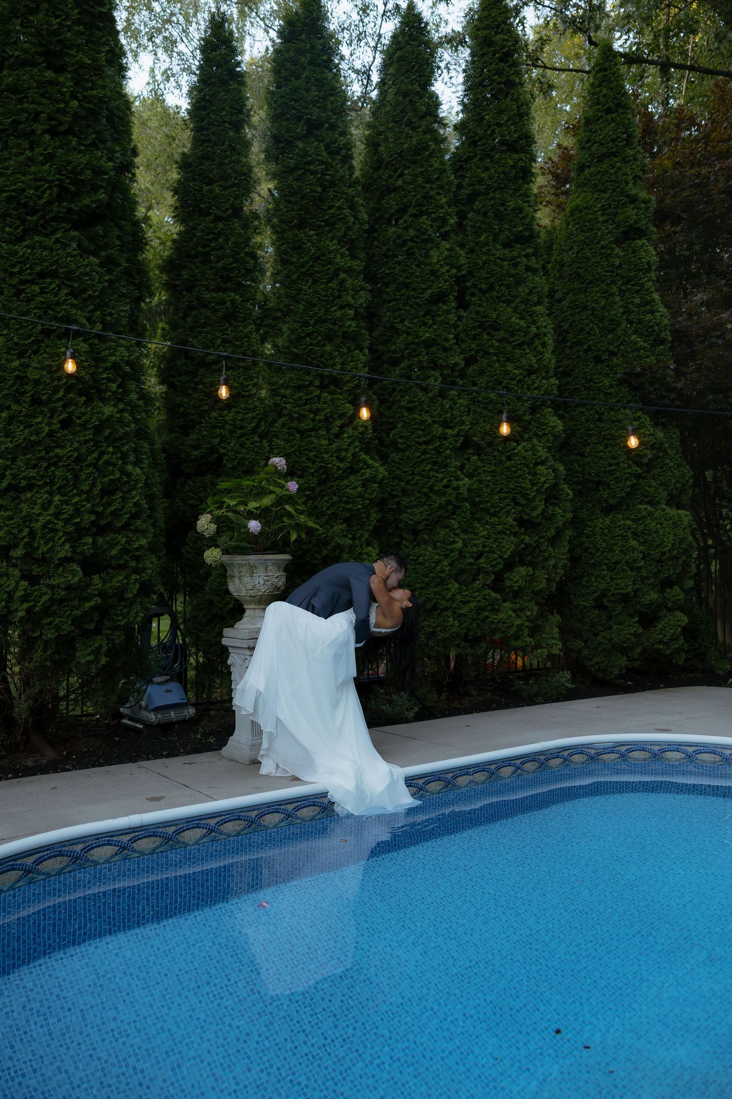 Bride and groom share a romantic dip kiss beside a pool, surrounded by tall evergreens and string lights at dusk.