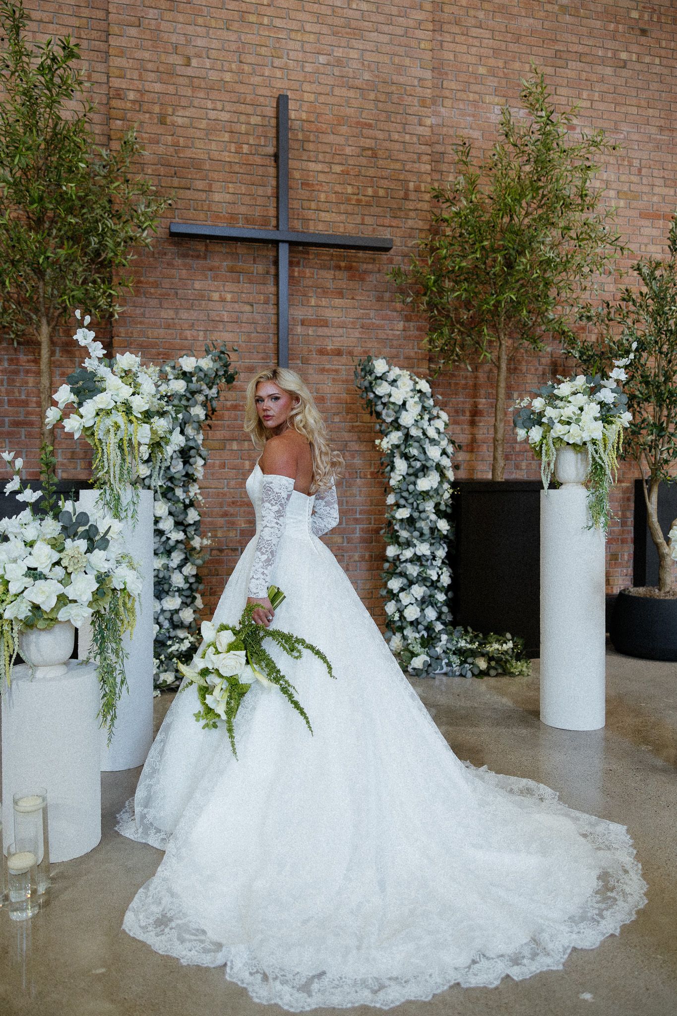 Bride in a white lace ball gown with a long train poses before a cross, surrounded by white floral arrangements.