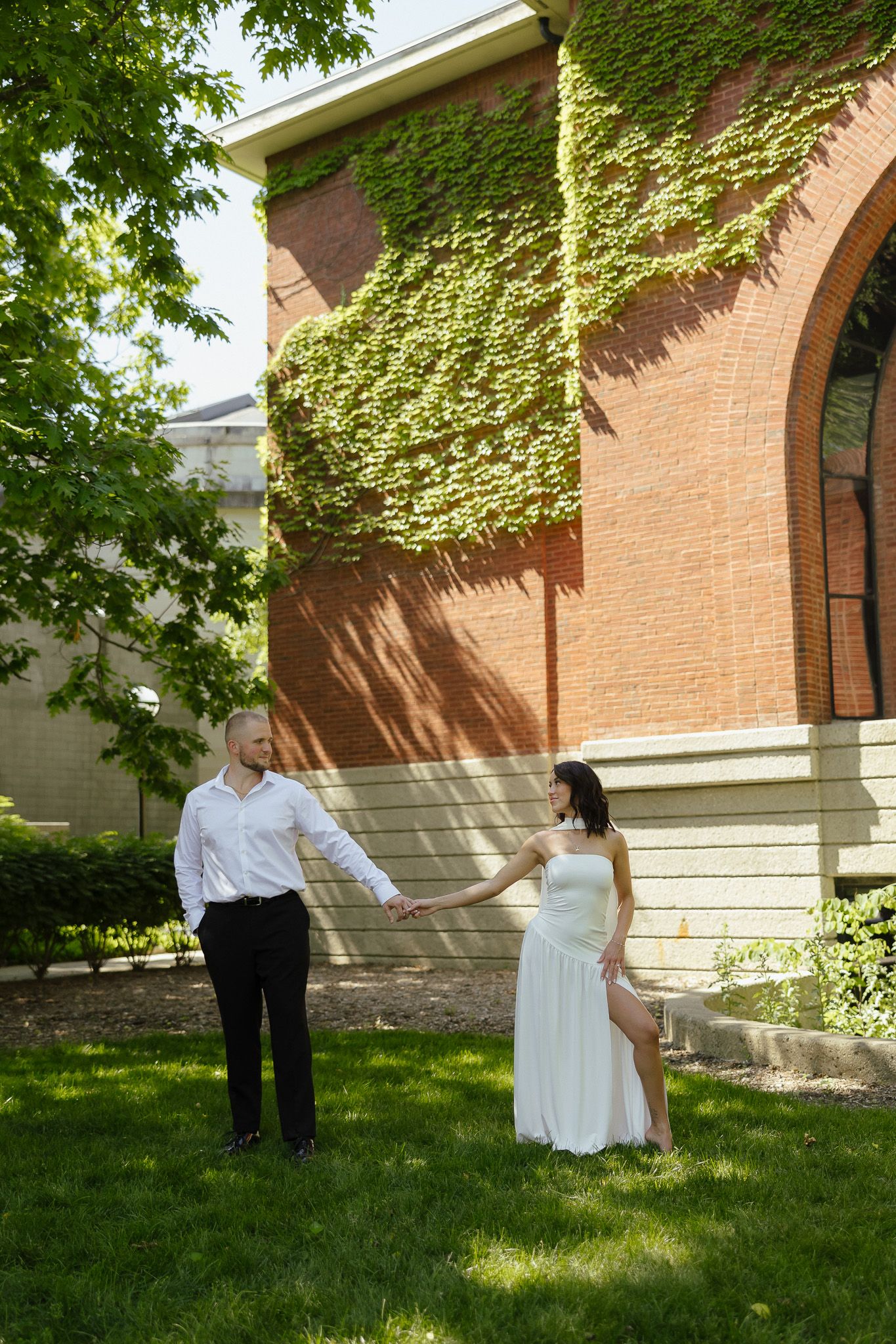 Couple holding hands on green lawn beside ivy-covered brick building on sunny day in white dress and suit.