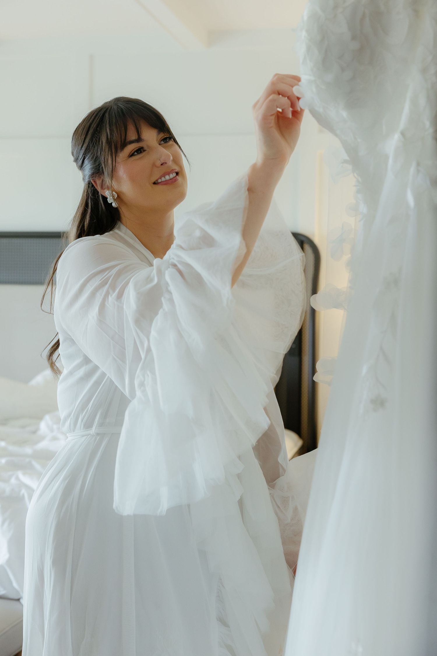 Smiling Asian bride in white robe touching her wedding dress hanging in a bright bedroom.