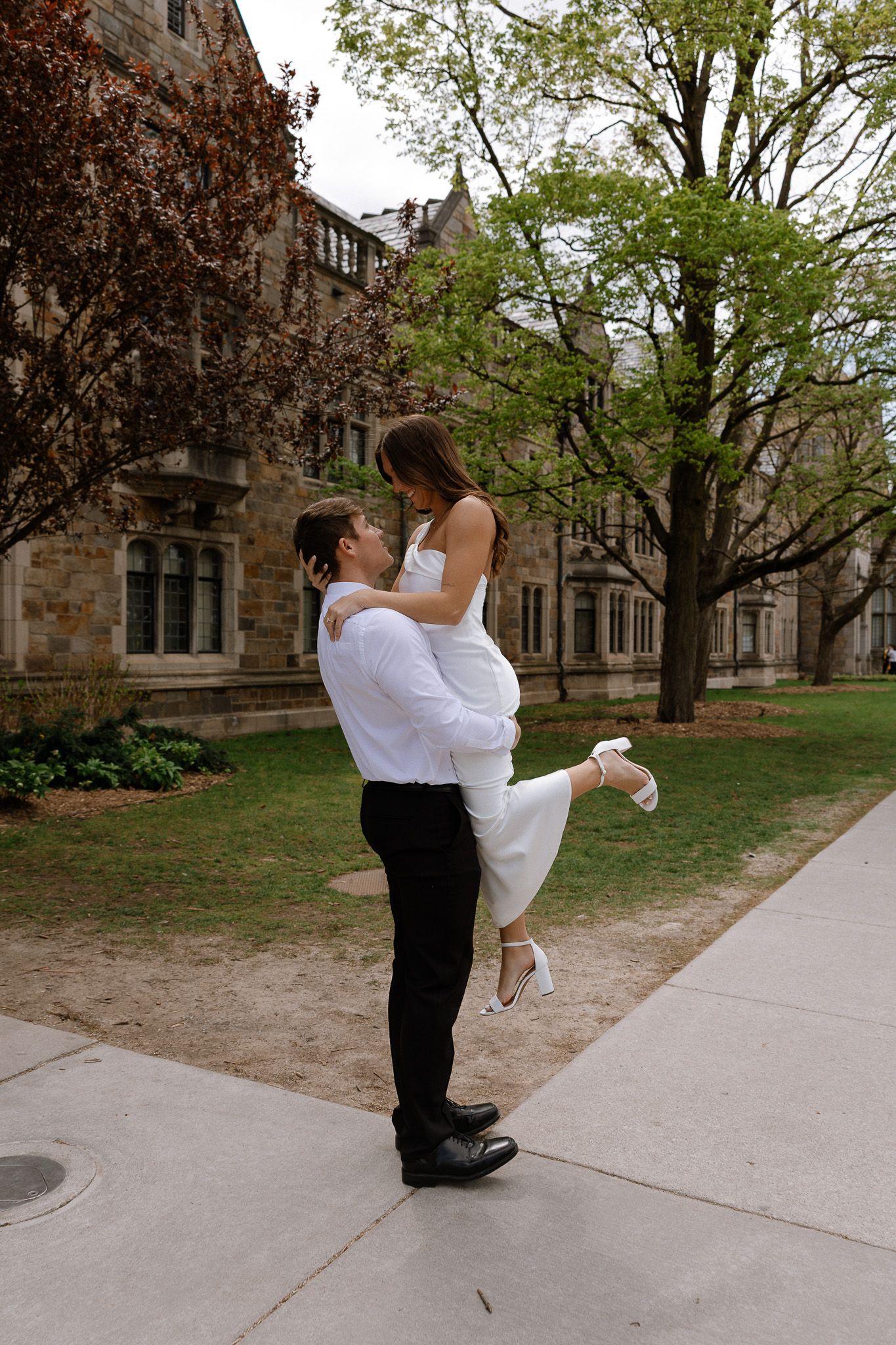 Groom lifting bride in white dress outdoors near historic stone building surrounded by lush green trees.