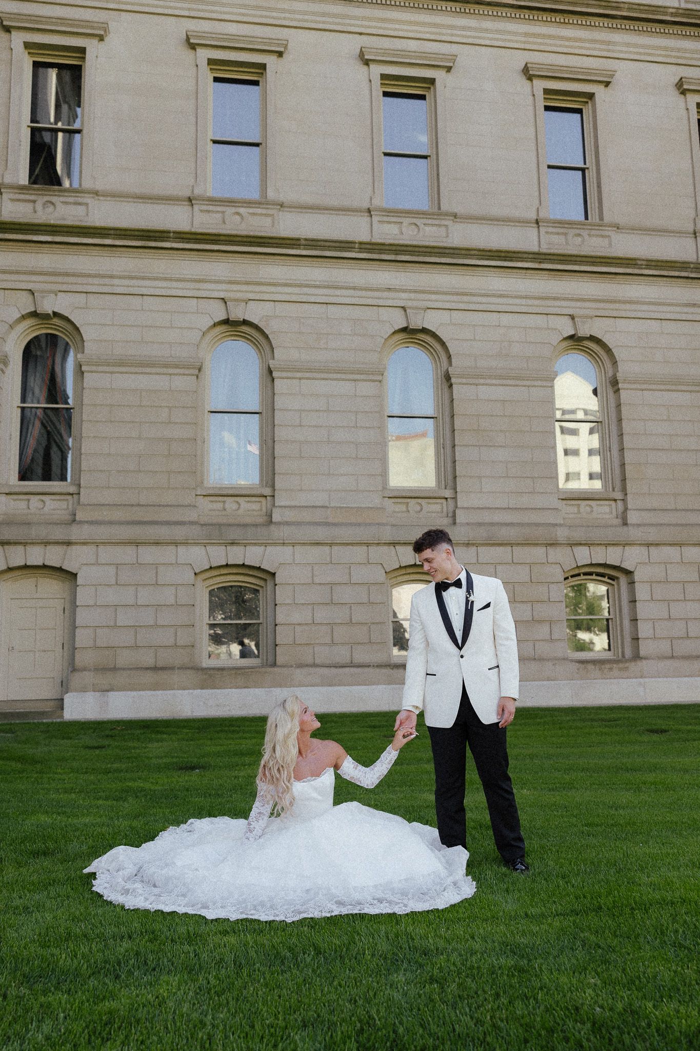Bride in white gown sits on green grass holding groom's hand in white tuxedo jacket outside stone building.