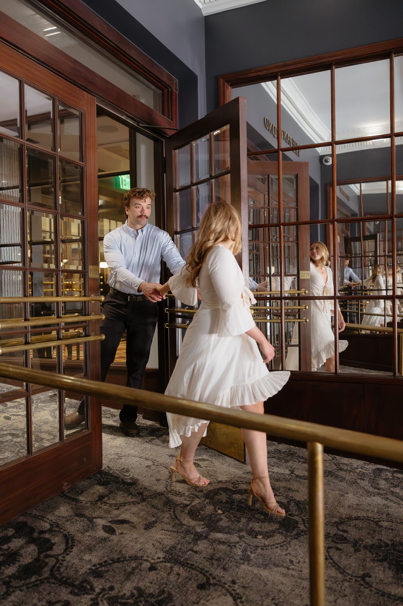 Man holding door open for woman in white dress entering elegant hotel lobby.