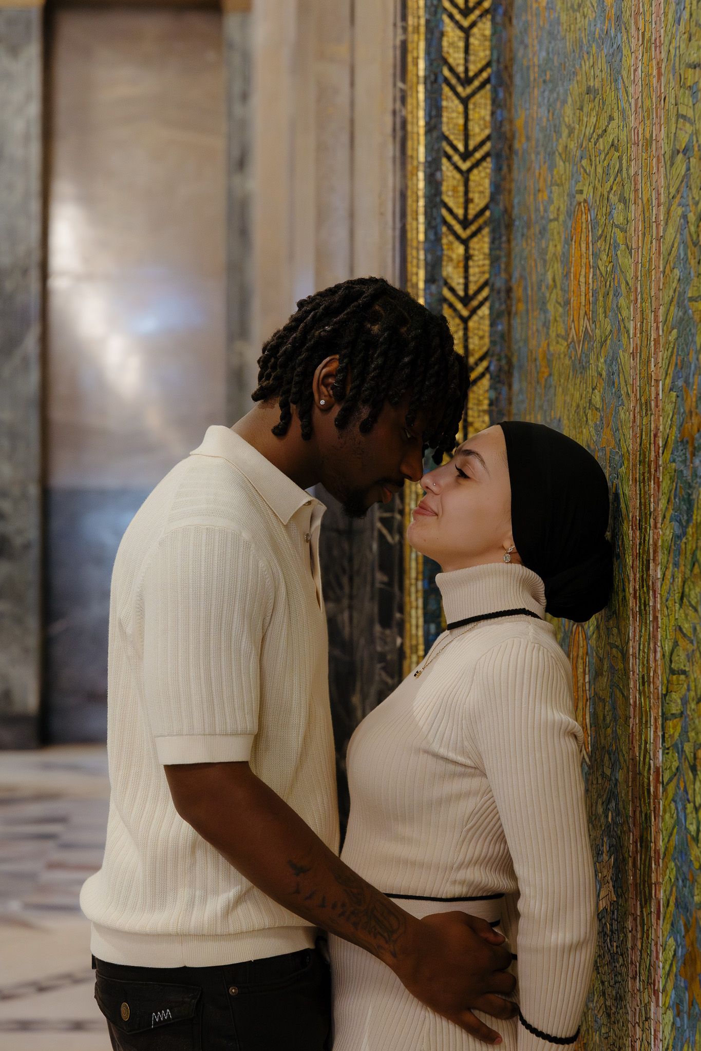 A couple sharing a romantic moment, leaning in for a kiss against a decorative mosaic wall in an elegant interior.