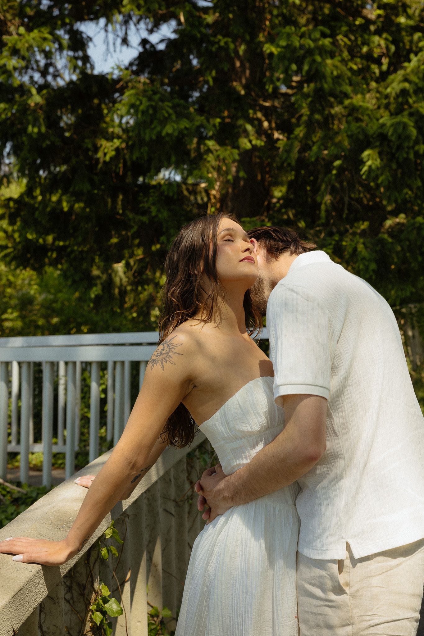 Romantic couple in white outfits sharing an intimate moment on a garden bridge surrounded by lush green trees.