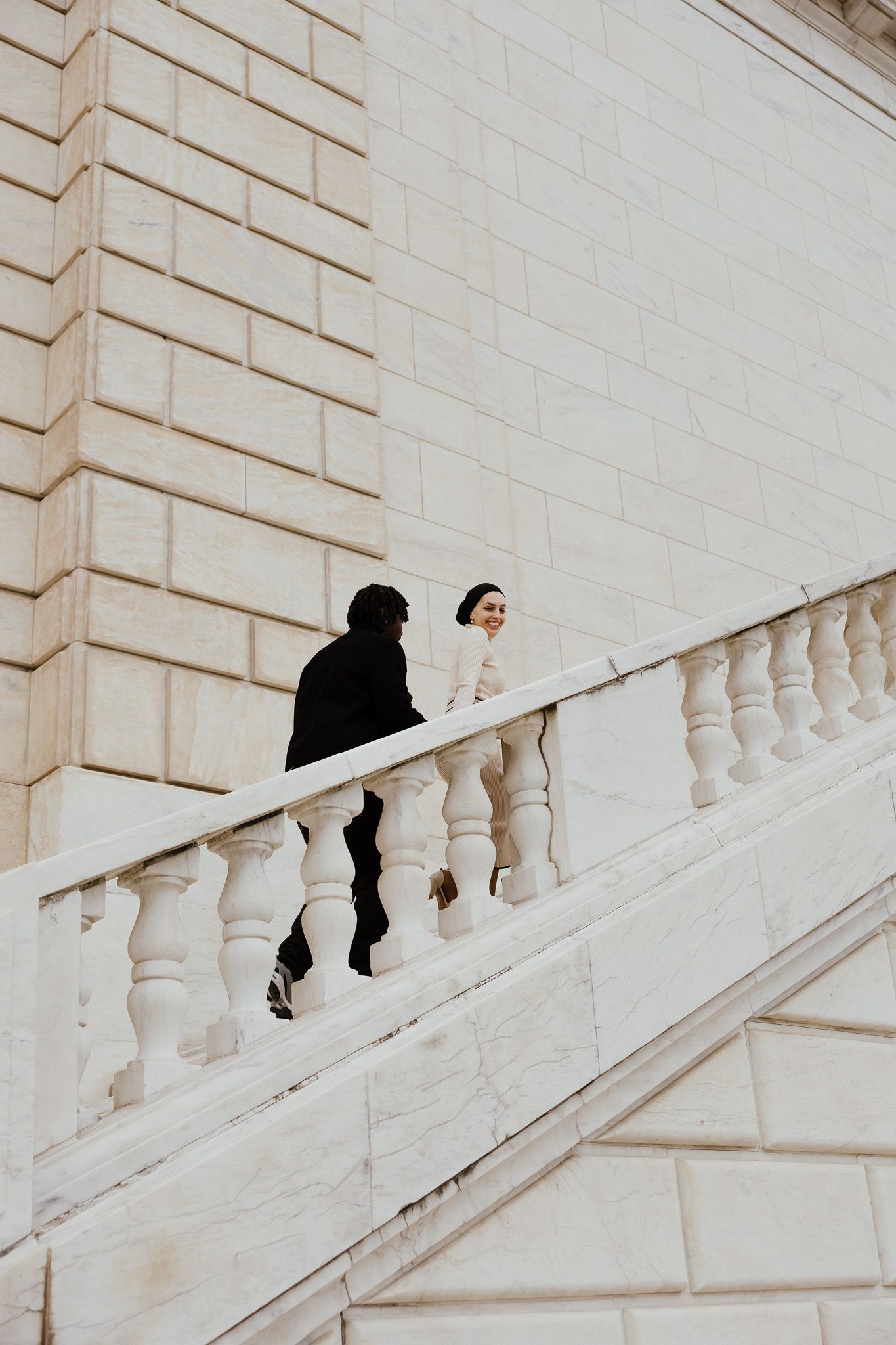 A couple walks up elegant white marble stairs with ornate balustrade at a grand neoclassical building.