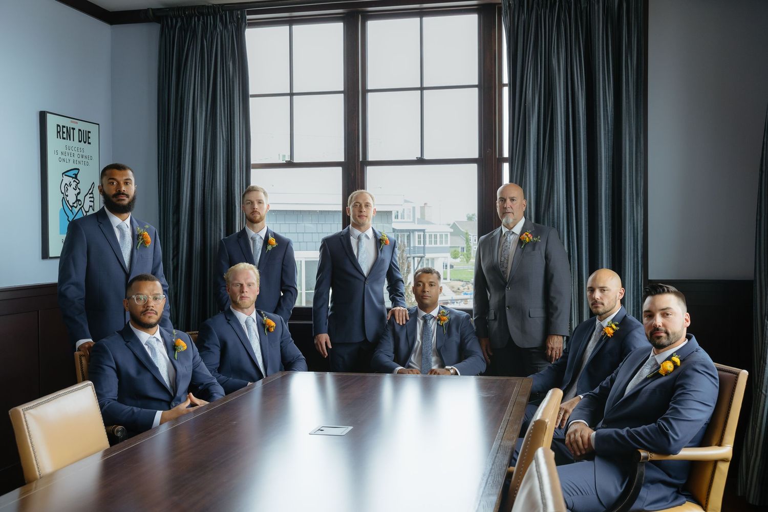 Group of nine groomsmen in navy suits with yellow boutonnieres posing in elegant boardroom with large windows.