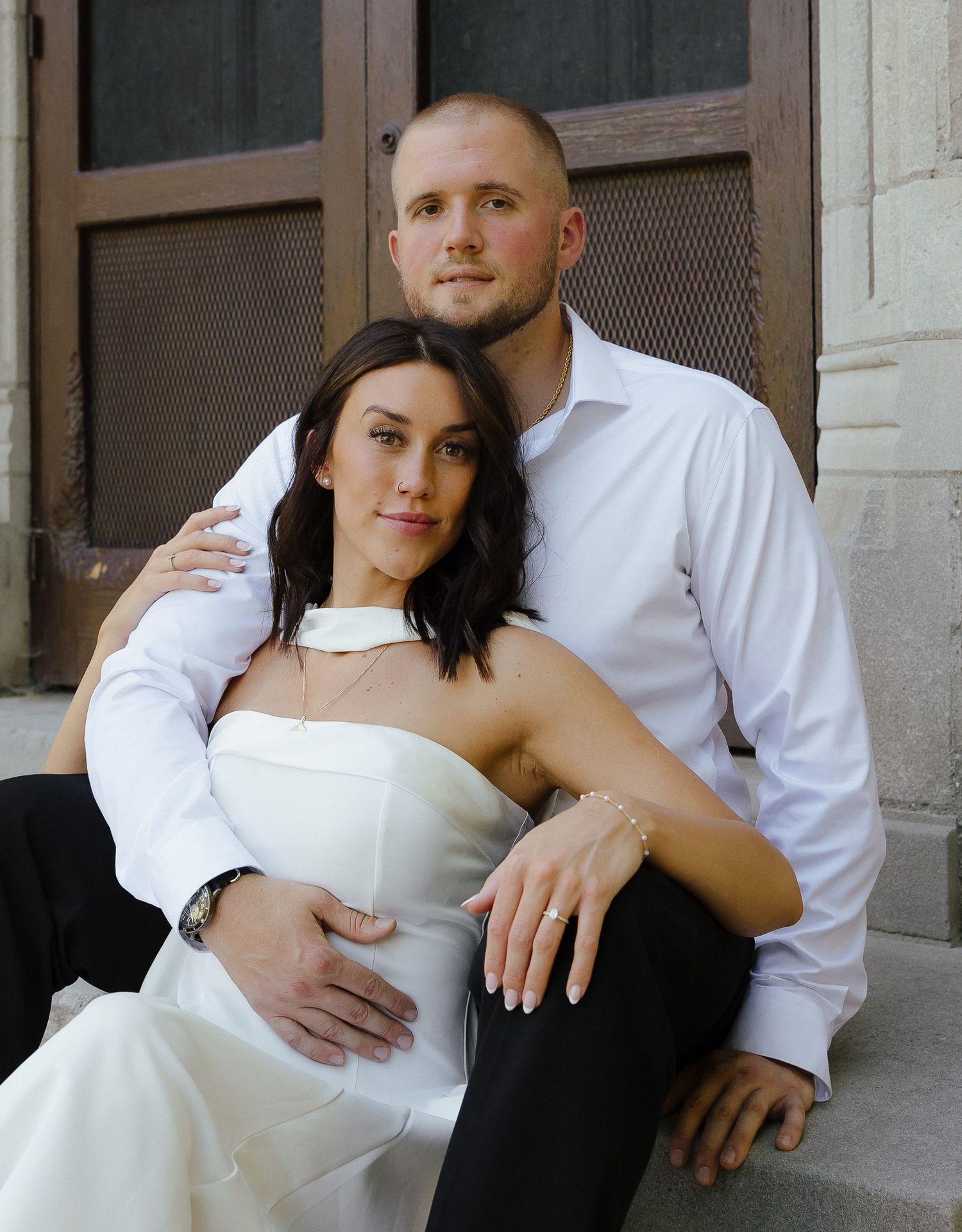 A couple in wedding attire sits on steps, the bride in white strapless gown leaning against the groom in a white dress shirt.